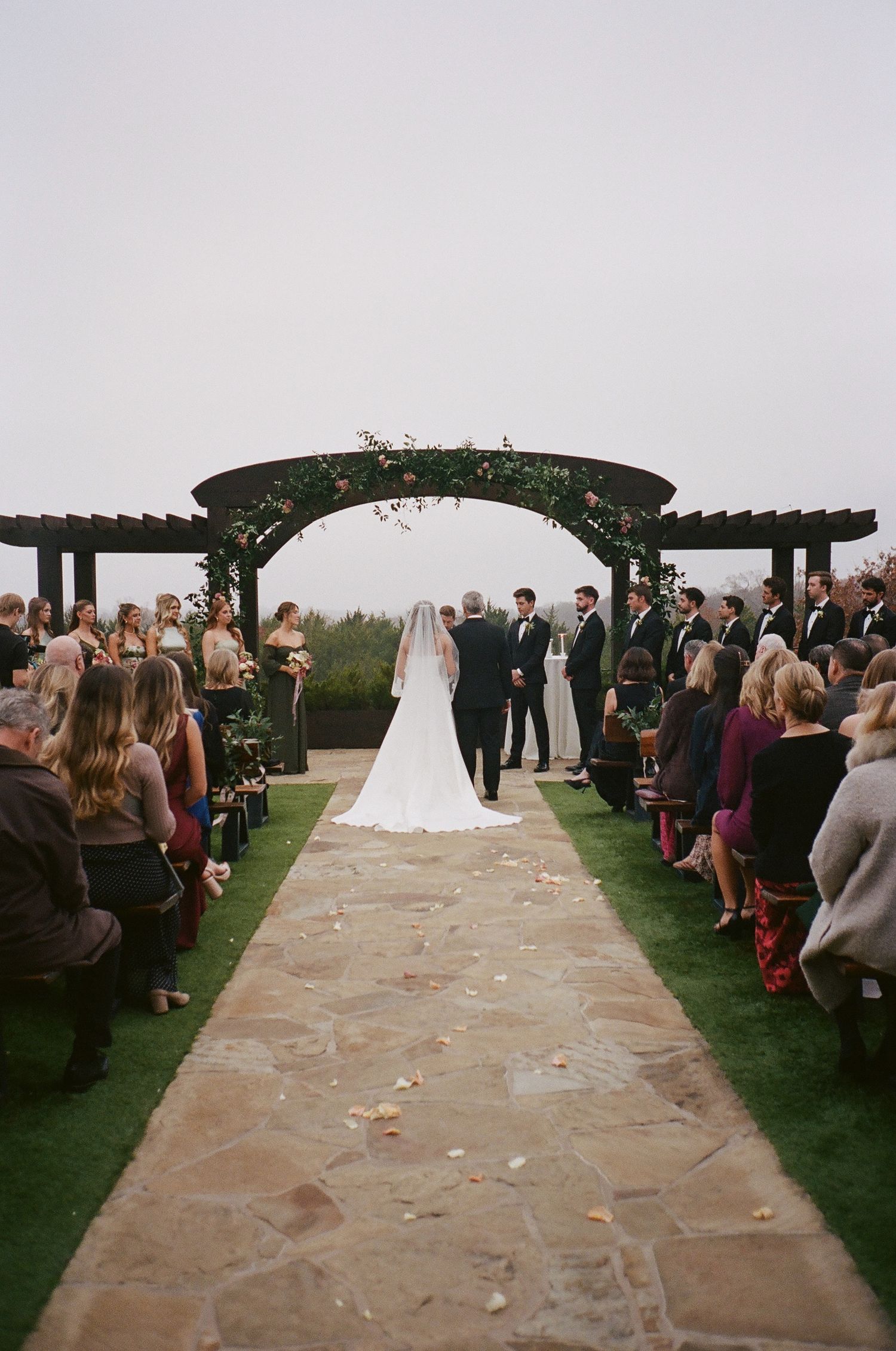 Outdoor wedding ceremony under dark pergola arch with stone aisle and guests.