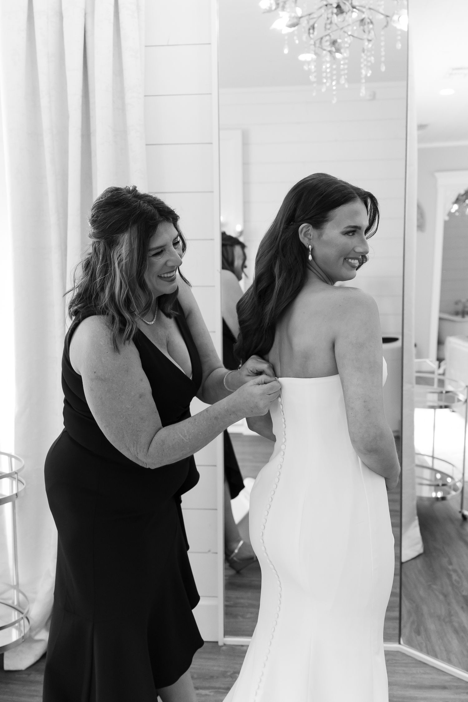 Black and white photo of woman helping bride button her strapless wedding gown in a bright dressing room.