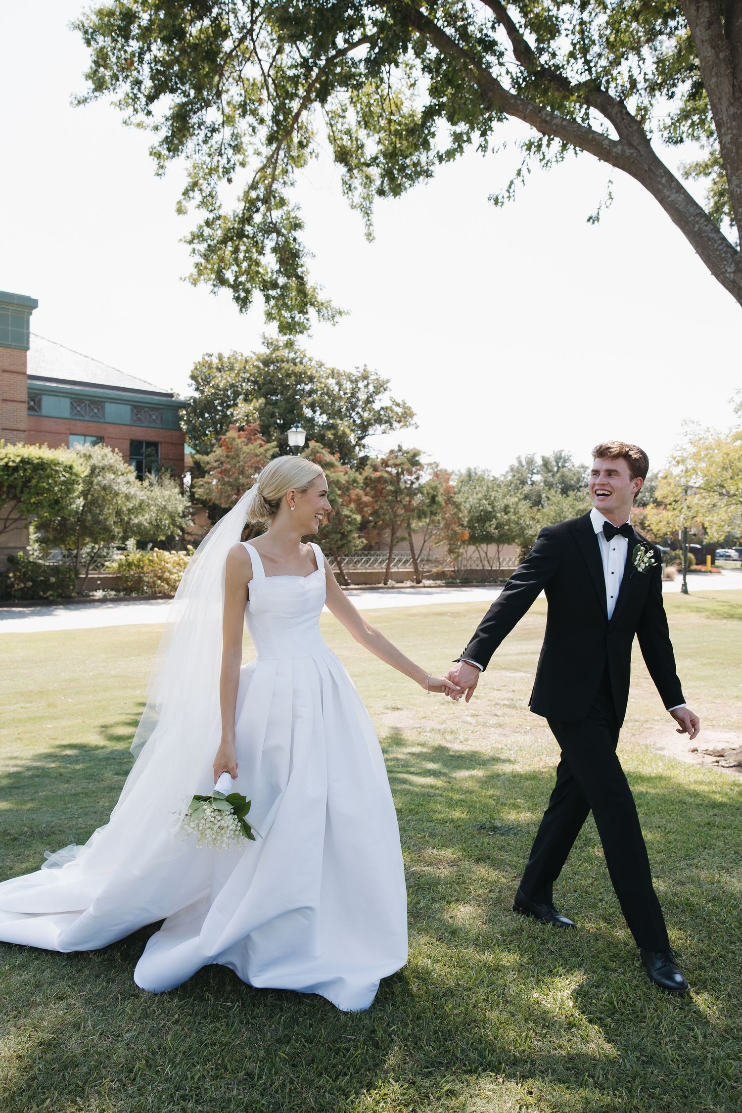 Bride and groom holding hands walking on green lawn, bride in white gown with veil, groom in black tuxedo.