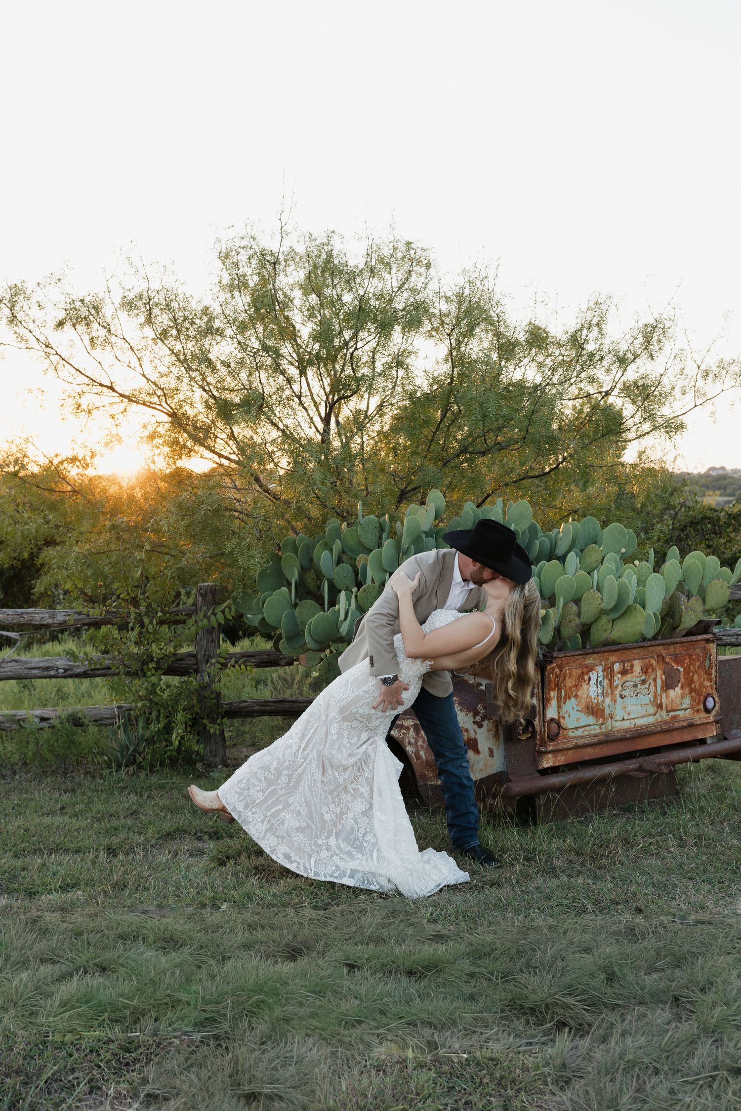 Groom in cowboy hat dips bride in lace gown at sunset near cactus and rustic fence.