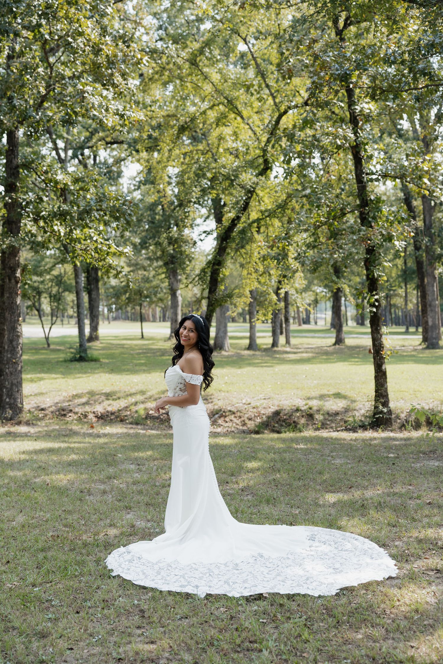Bride in off-shoulder white gown with lace train poses in a lush green wooded park.