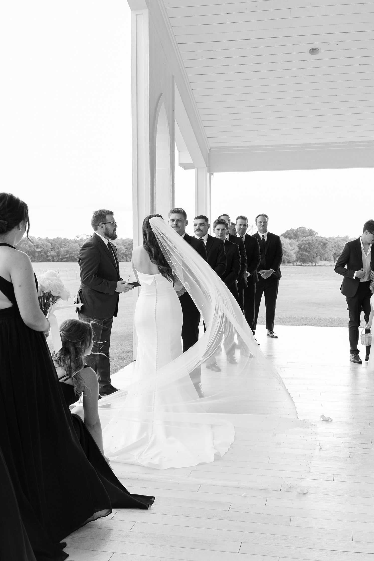 Black and white photo of bride in cathedral veil at outdoor wedding ceremony with wedding party.