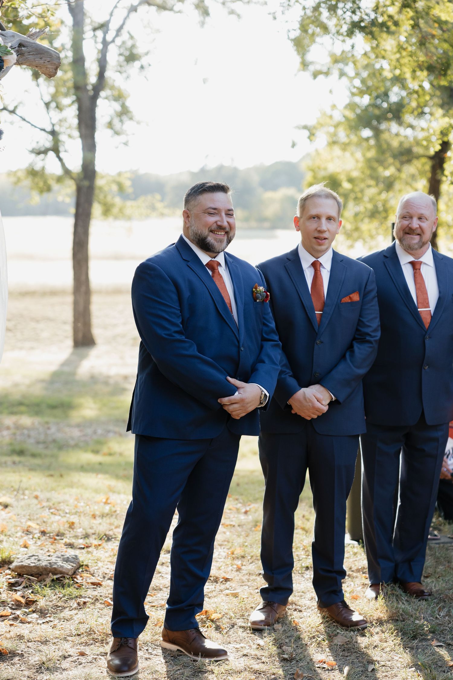 Groom and groomsmen in navy suits with rust ties smiling outdoors at wedding ceremony.