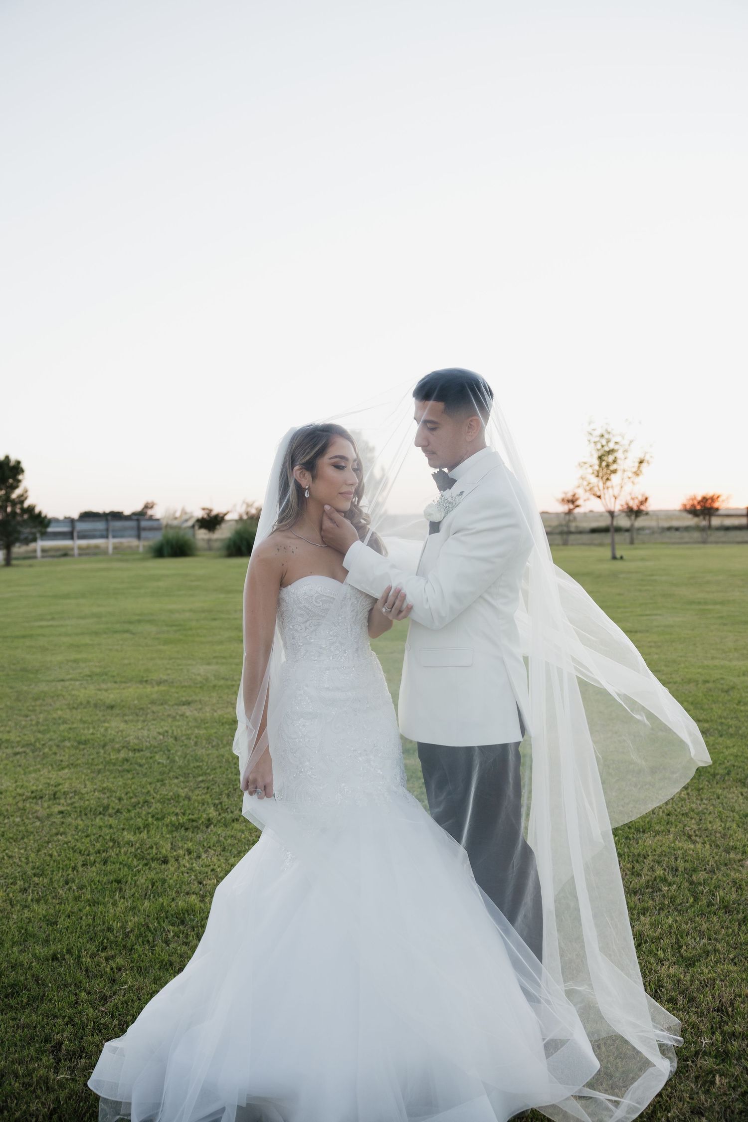 Bride and groom stand beneath flowing veil at sunset on green lawn in romantic portrait.