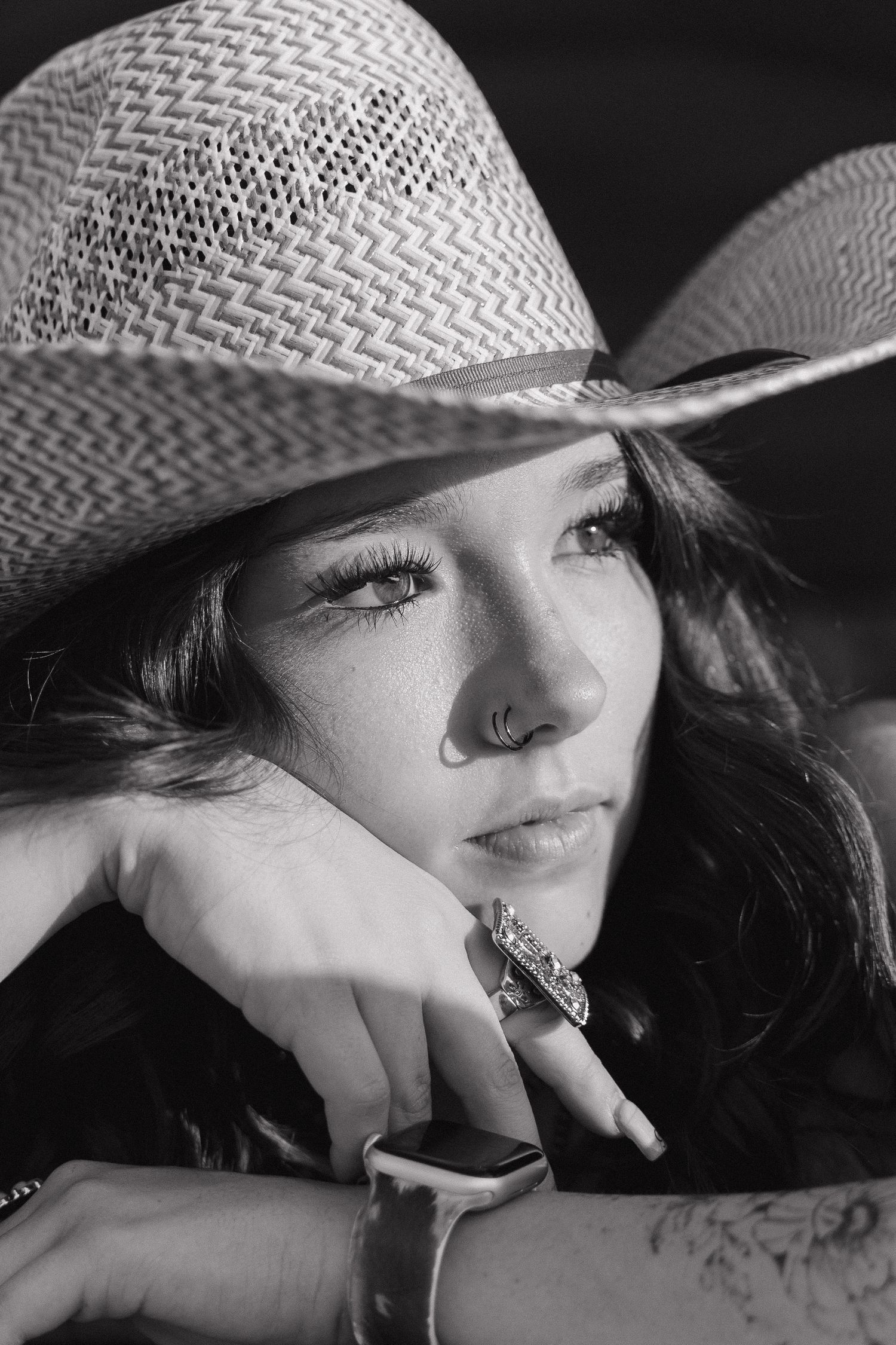 Black and white portrait of a young woman wearing a straw cowboy hat, nose ring, and rings on her fingers.