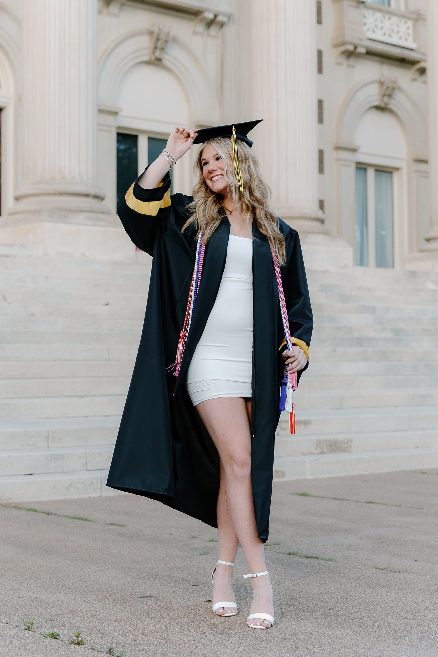 Smiling graduate in black cap and gown over white mini dress with colorful honor cords on university steps.