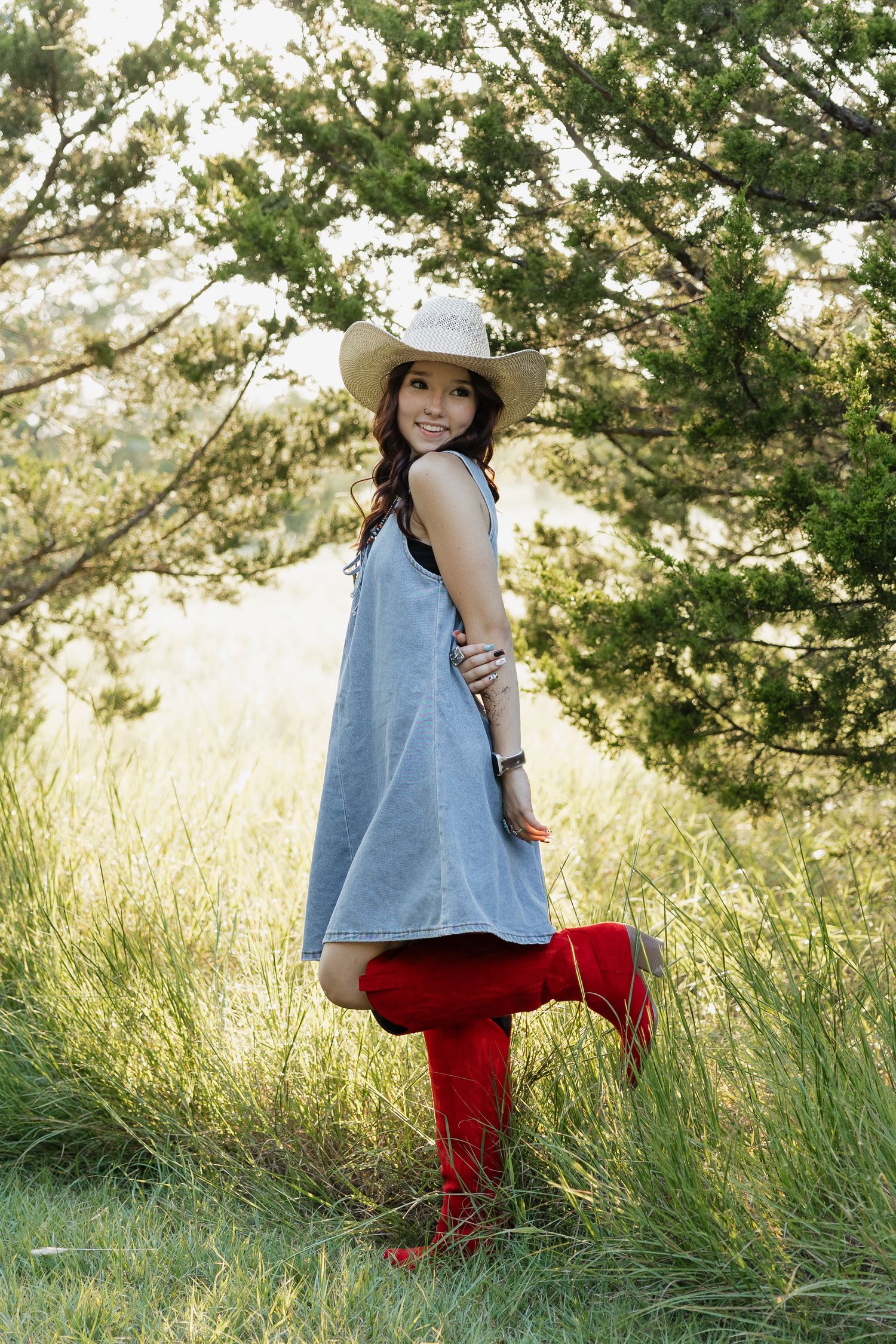 Young woman in denim dress and red cowboy boots posing sideways in a lush green field wearing a straw hat.