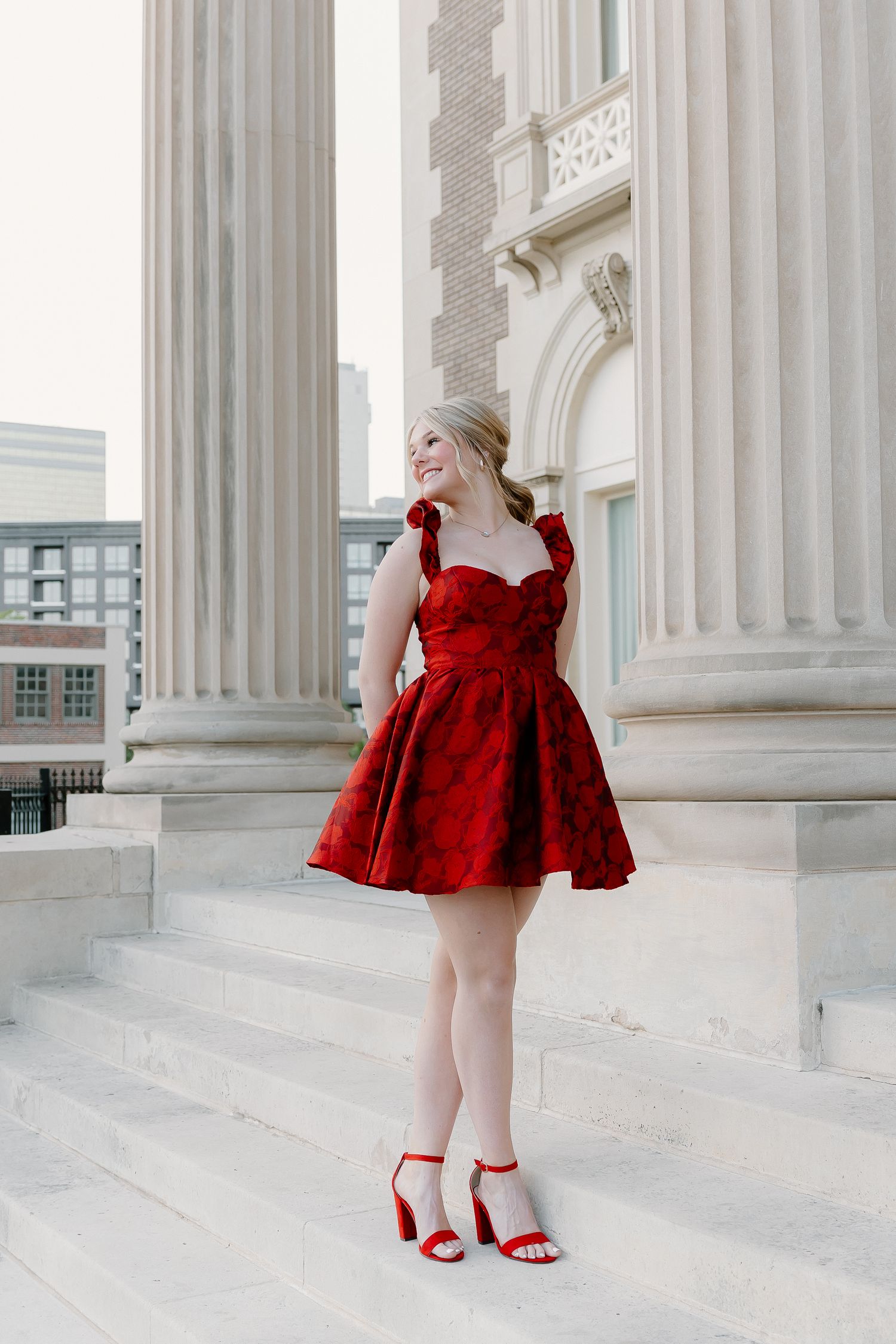 Blonde woman in elegant red floral sweetheart dress with red heeled sandals standing on marble steps by columns.