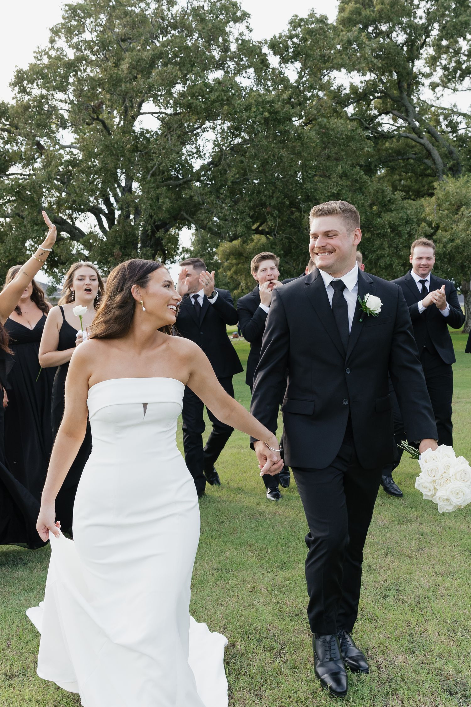 Newlyweds walk hand in hand as cheering wedding party celebrates behind them on green lawn.