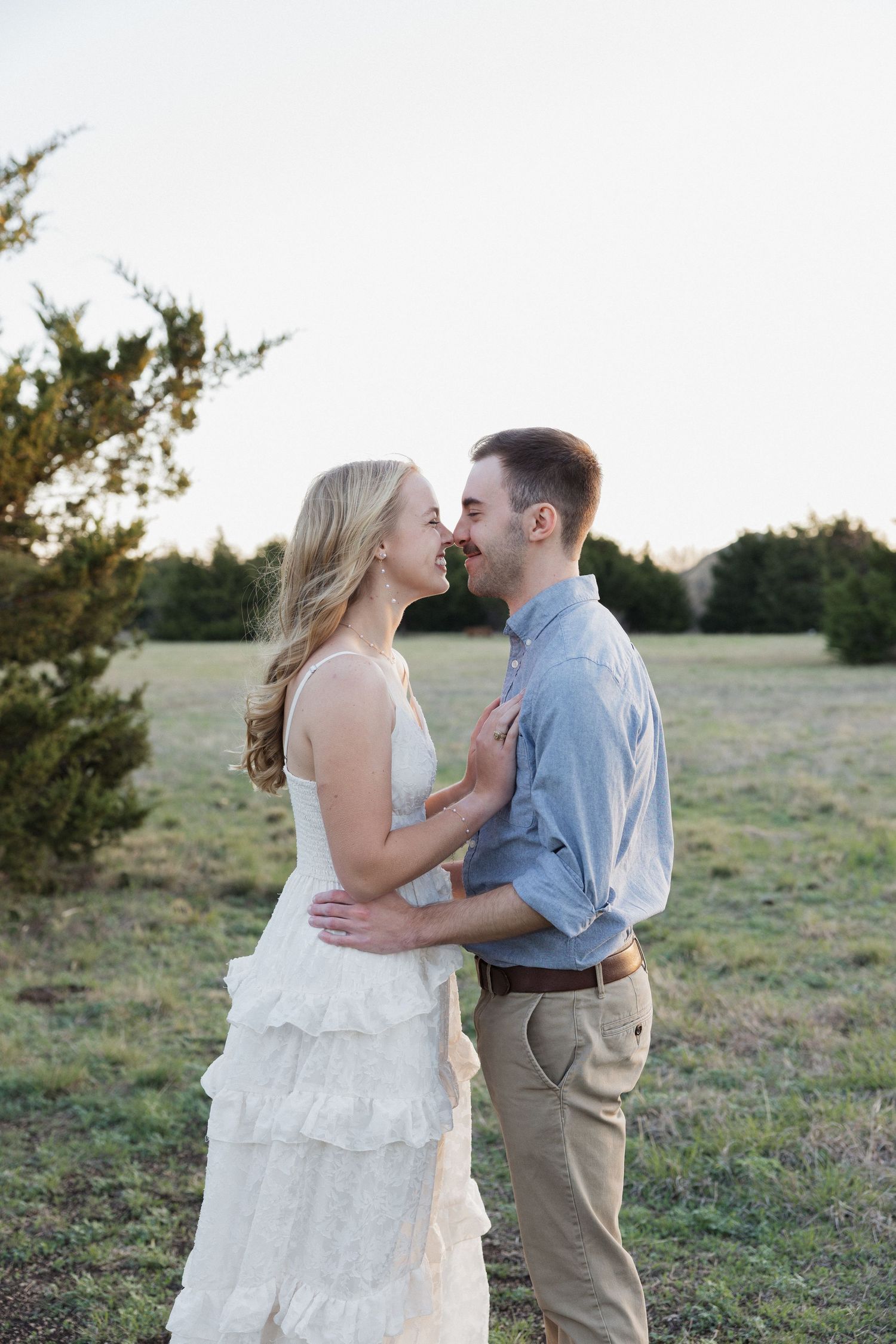A couple sharing a romantic moment in a field, woman in white ruffled dress, man in blue shirt and khakis.