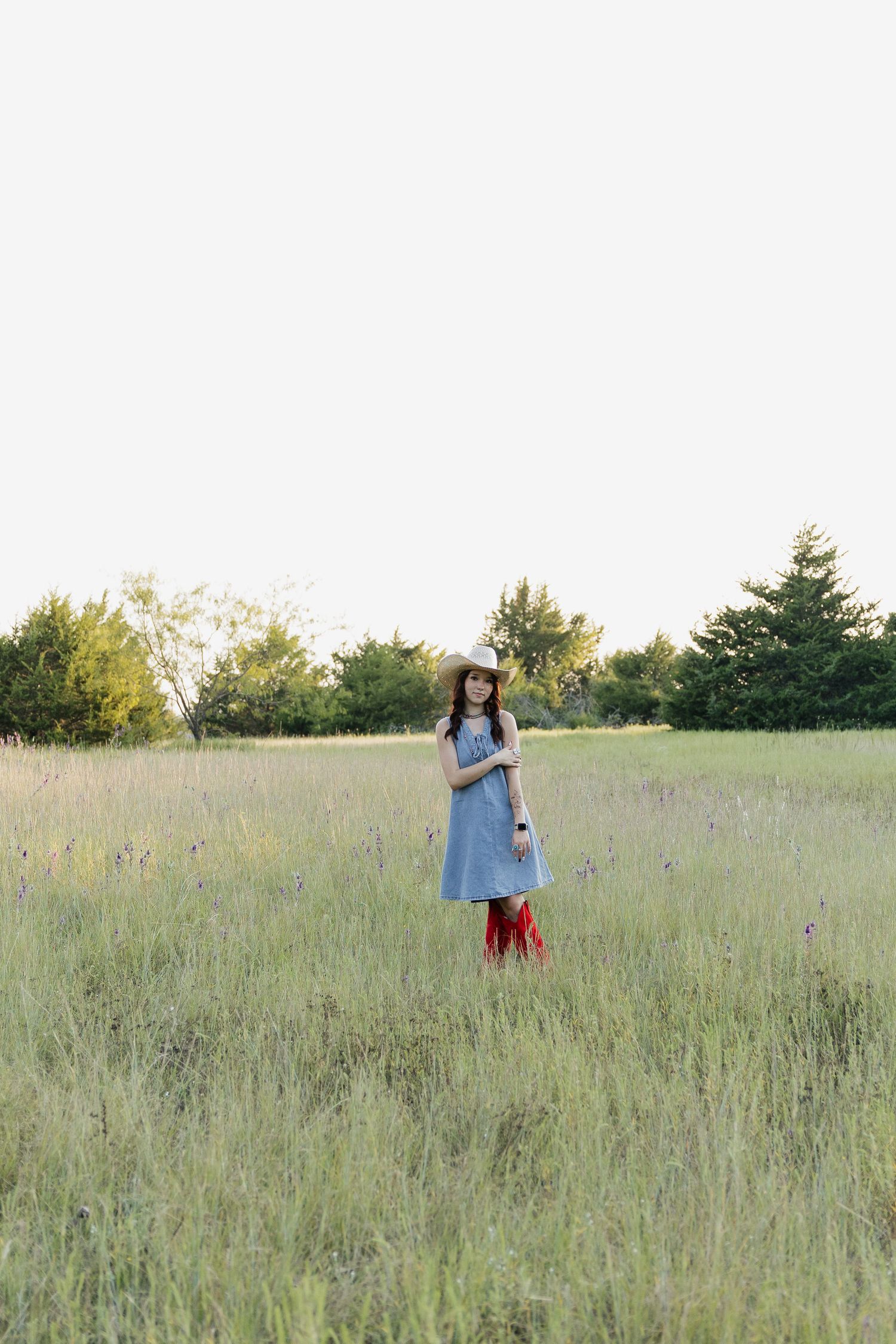 Young woman in a denim dress, straw hat, and red cowboy boots standing alone in a wide open grassy field.