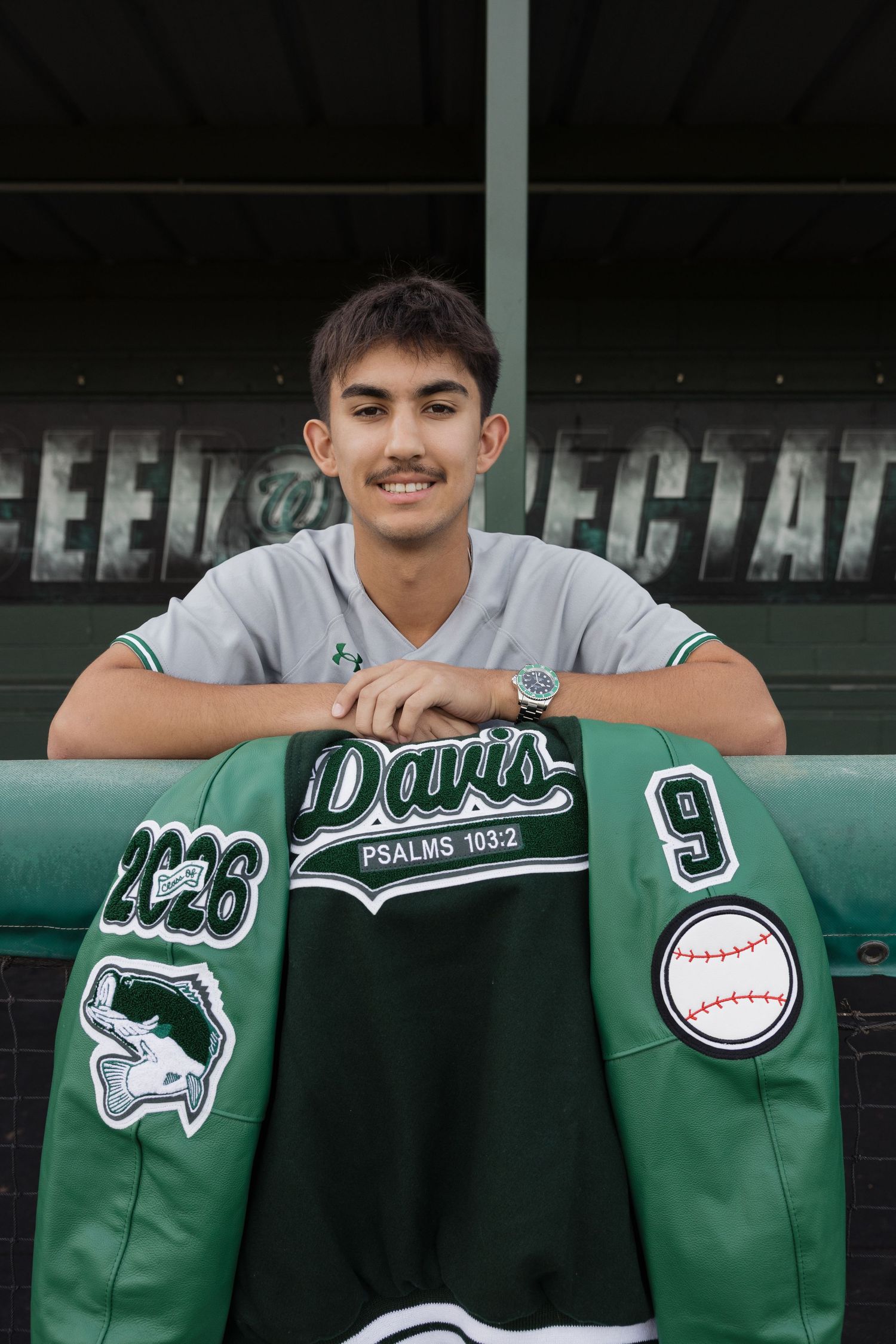 A smiling teen poses with a green Davis baseball letterman jacket numbered 9, sitting in a dugout wearing a gray jersey.