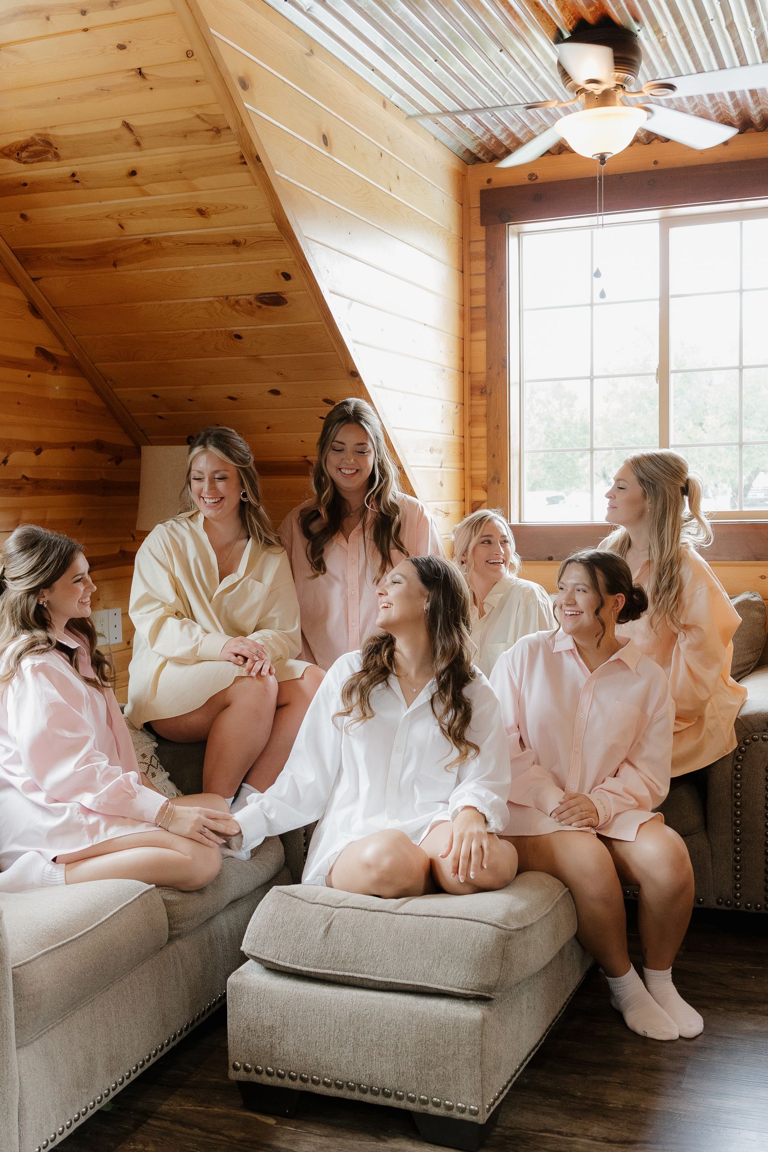 Bride in white robe sits surrounded by bridesmaids in pink and cream satin robes inside a rustic wooden cabin.