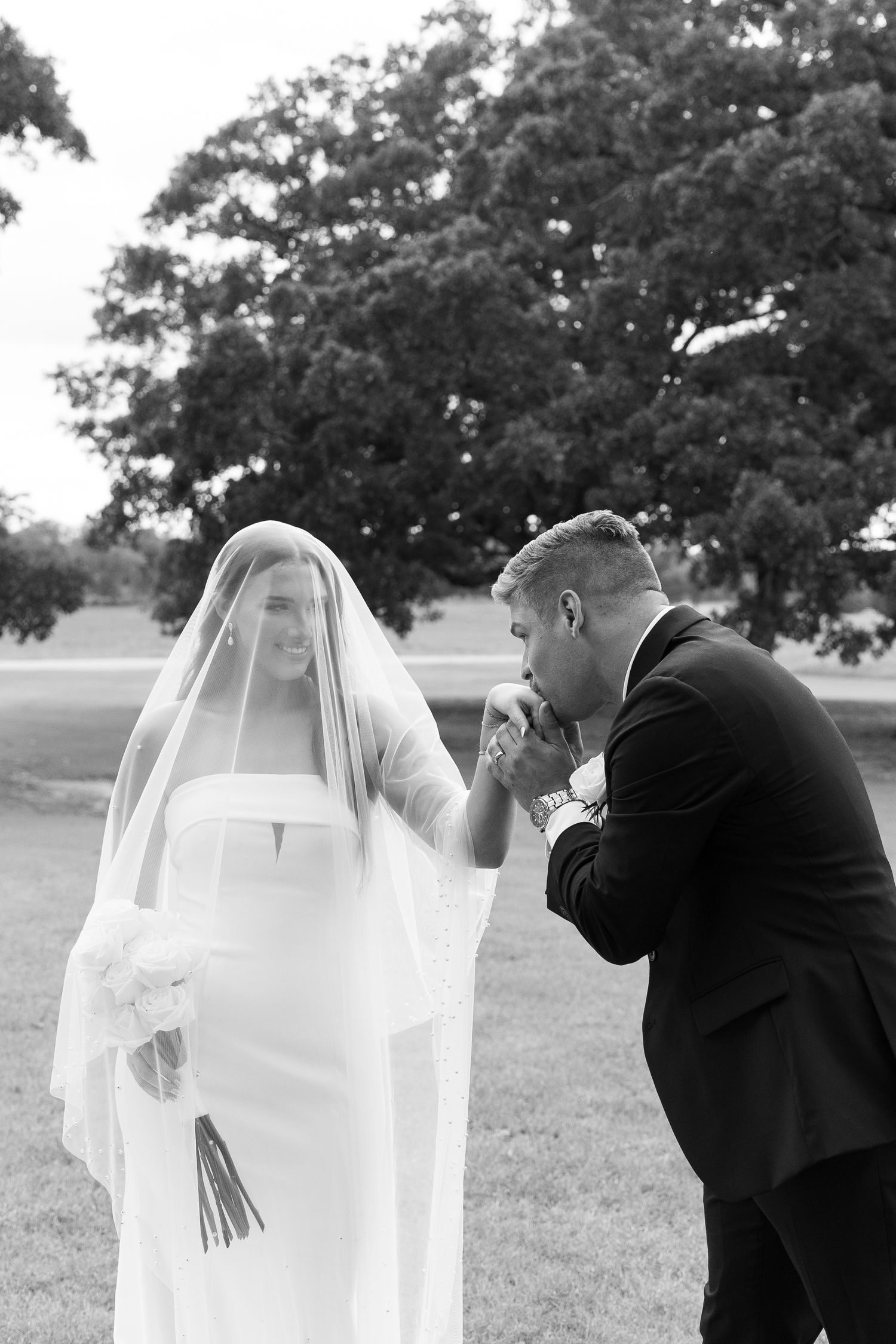 Black and white photo of groom kissing brides hand during romantic first look under wedding veil.