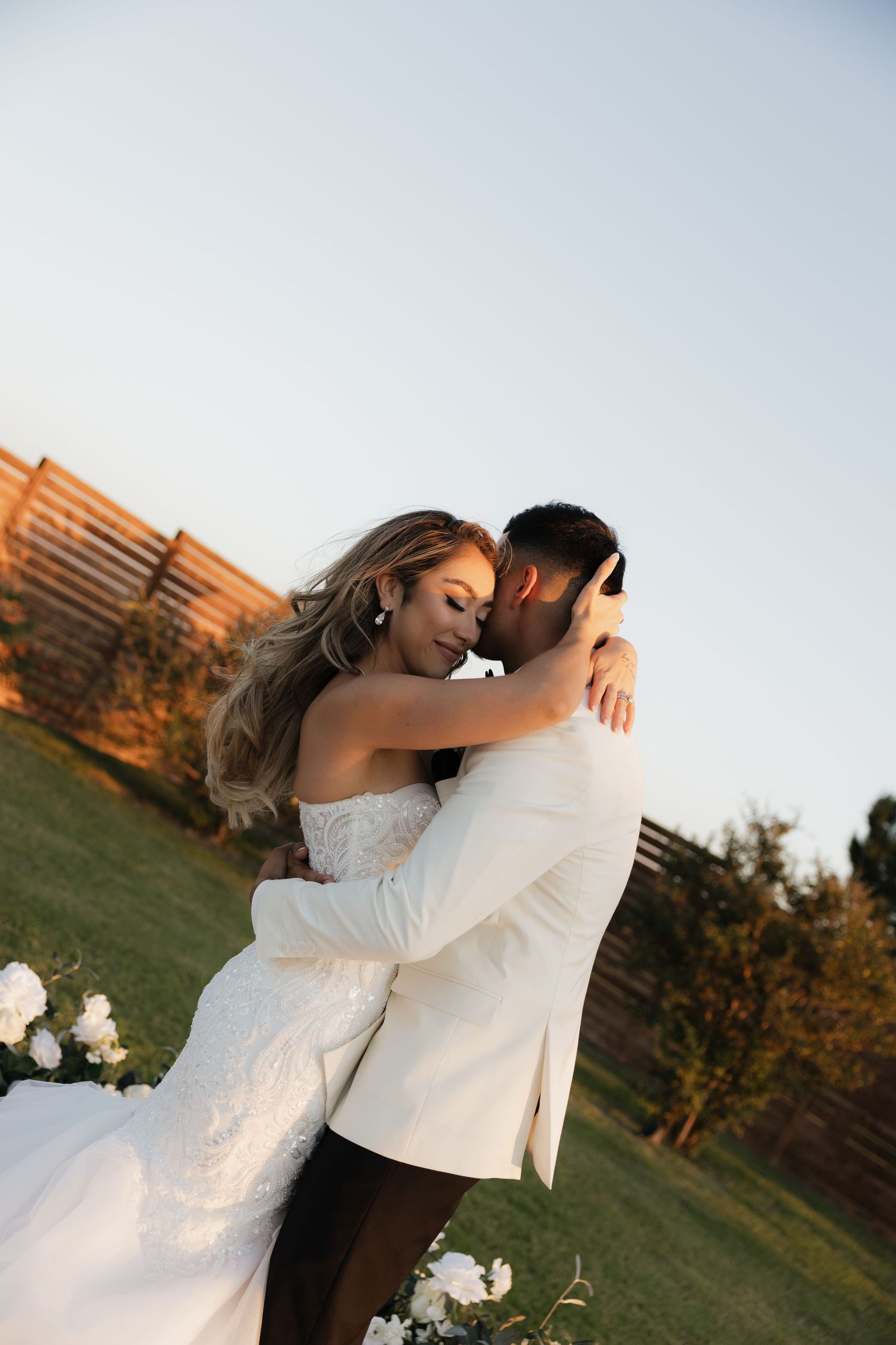 Bride and groom embrace tenderly in golden hour light beside white floral wedding arch outdoors.