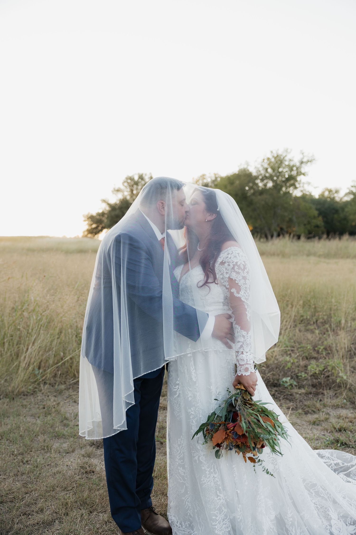 Bride and groom share a romantic kiss under flowing veil in a golden field at sunset.