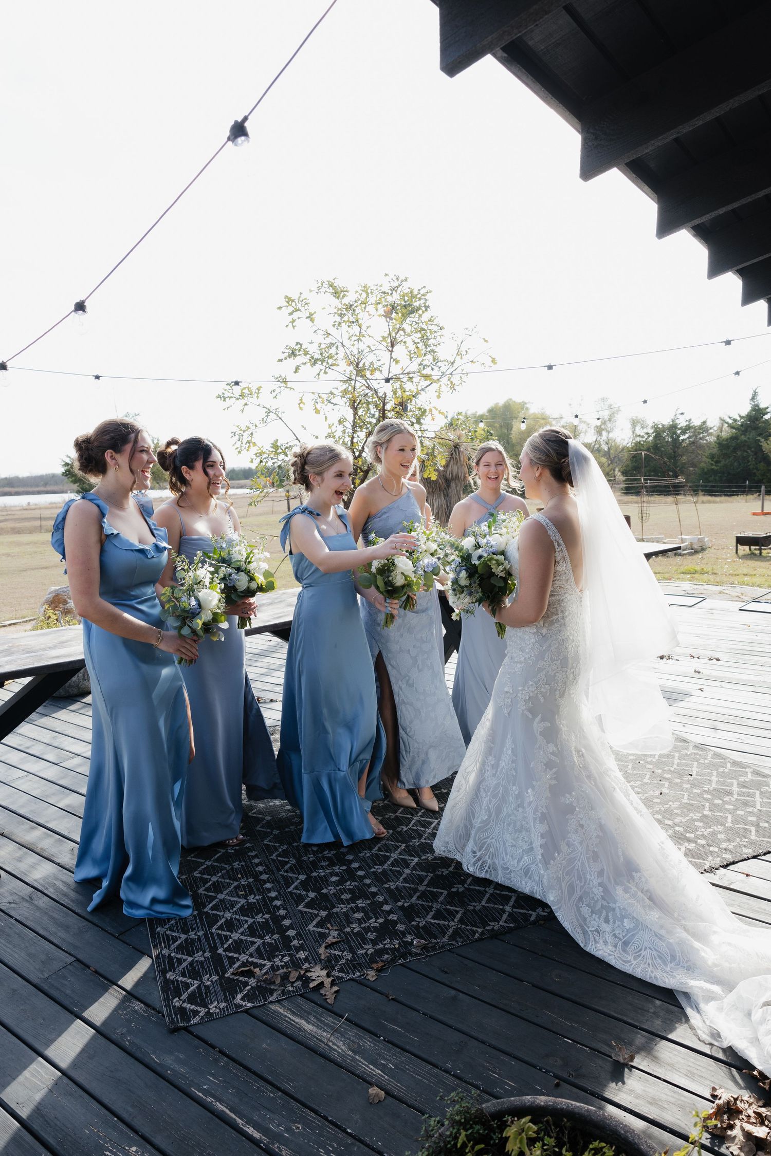 Bridesmaids in dusty blue gowns reacting with joy as bride reveals her lace wedding dress on deck.