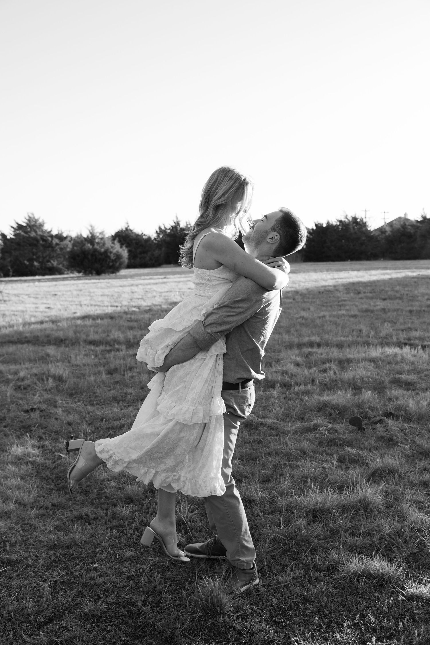 Black and white photo of a man lifting a woman in a white dress, sharing a romantic moment in an open field.