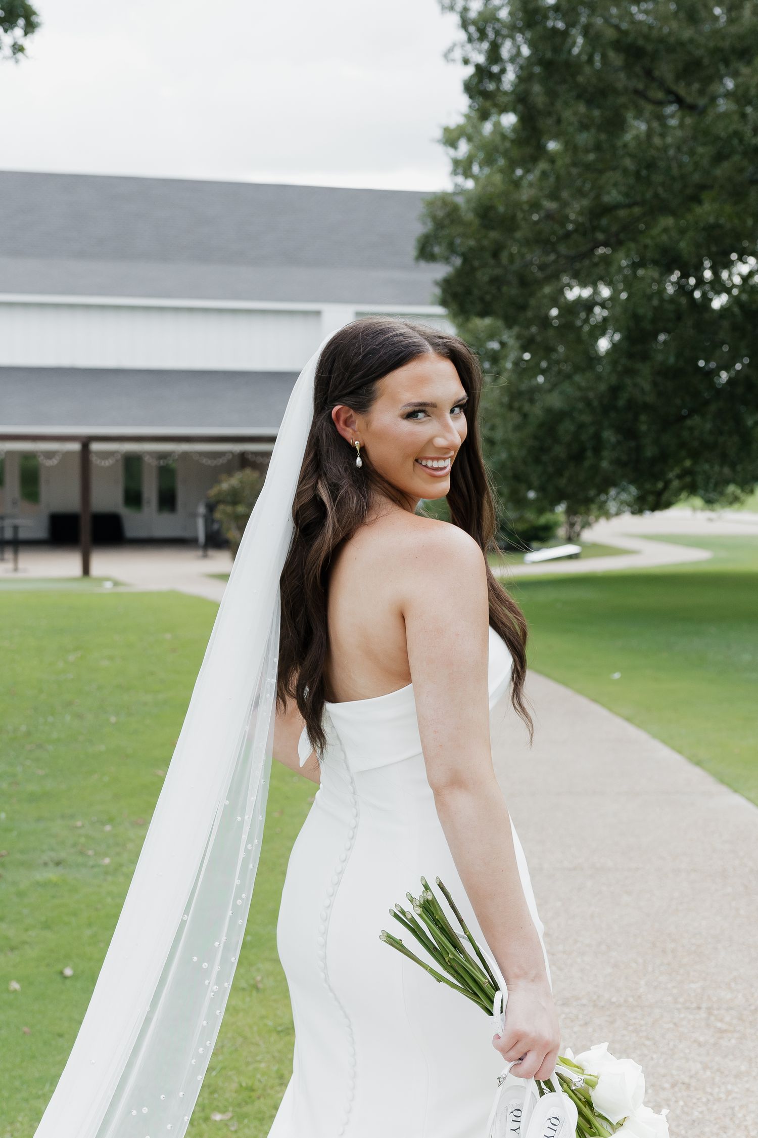Bride in strapless white gown and cathedral veil smiles over shoulder holding white bouquet outdoors.