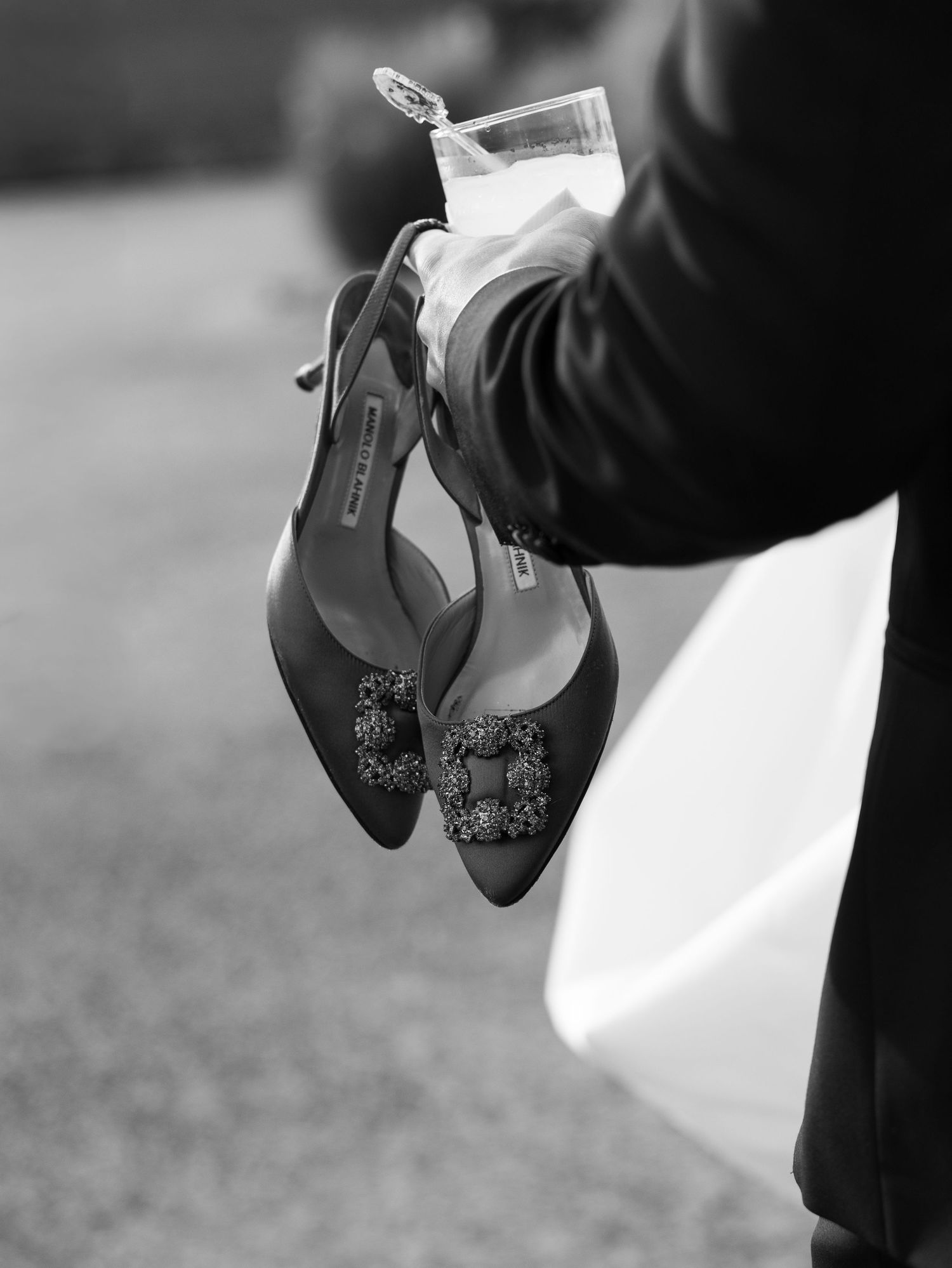 Black and white close-up of Manolo Blahnik jeweled heels being held alongside a champagne glass.