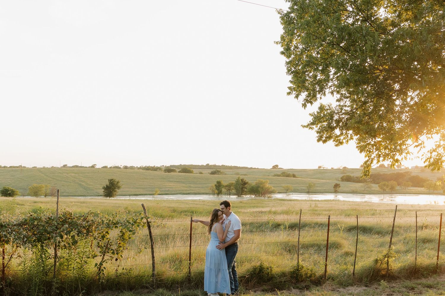 Couple embracing at golden hour sunset near rustic fence with open field and pond in background.