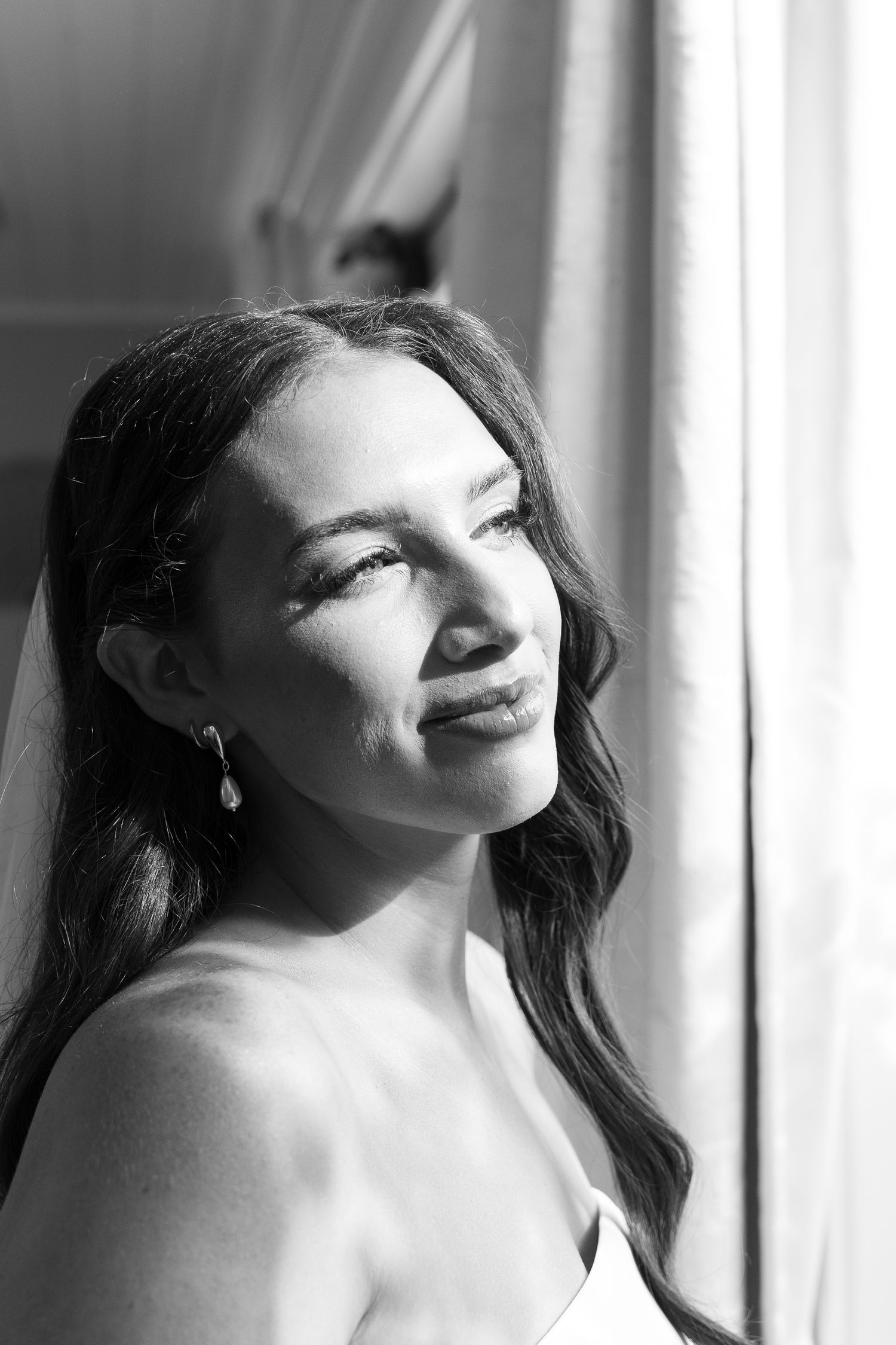 Black and white portrait of a bride with veil and drop earrings smiling softly by a window.