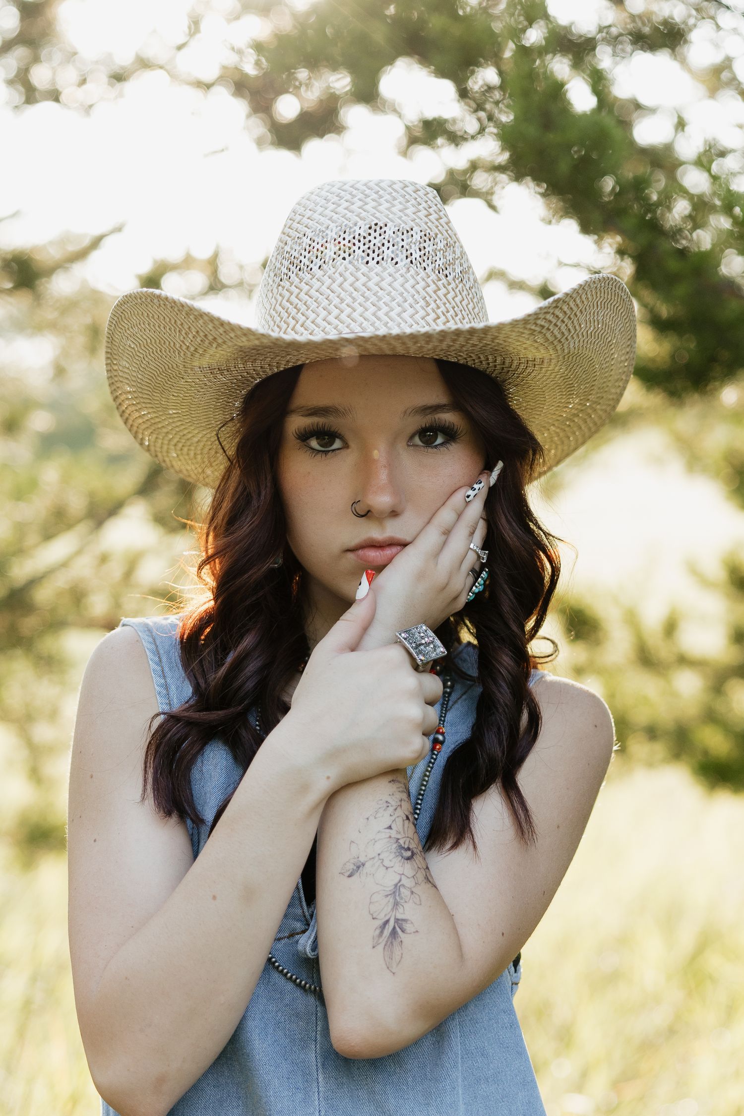 Young woman with dark wavy hair wearing a straw cowboy hat and denim top with a floral hand tattoo outdoors.