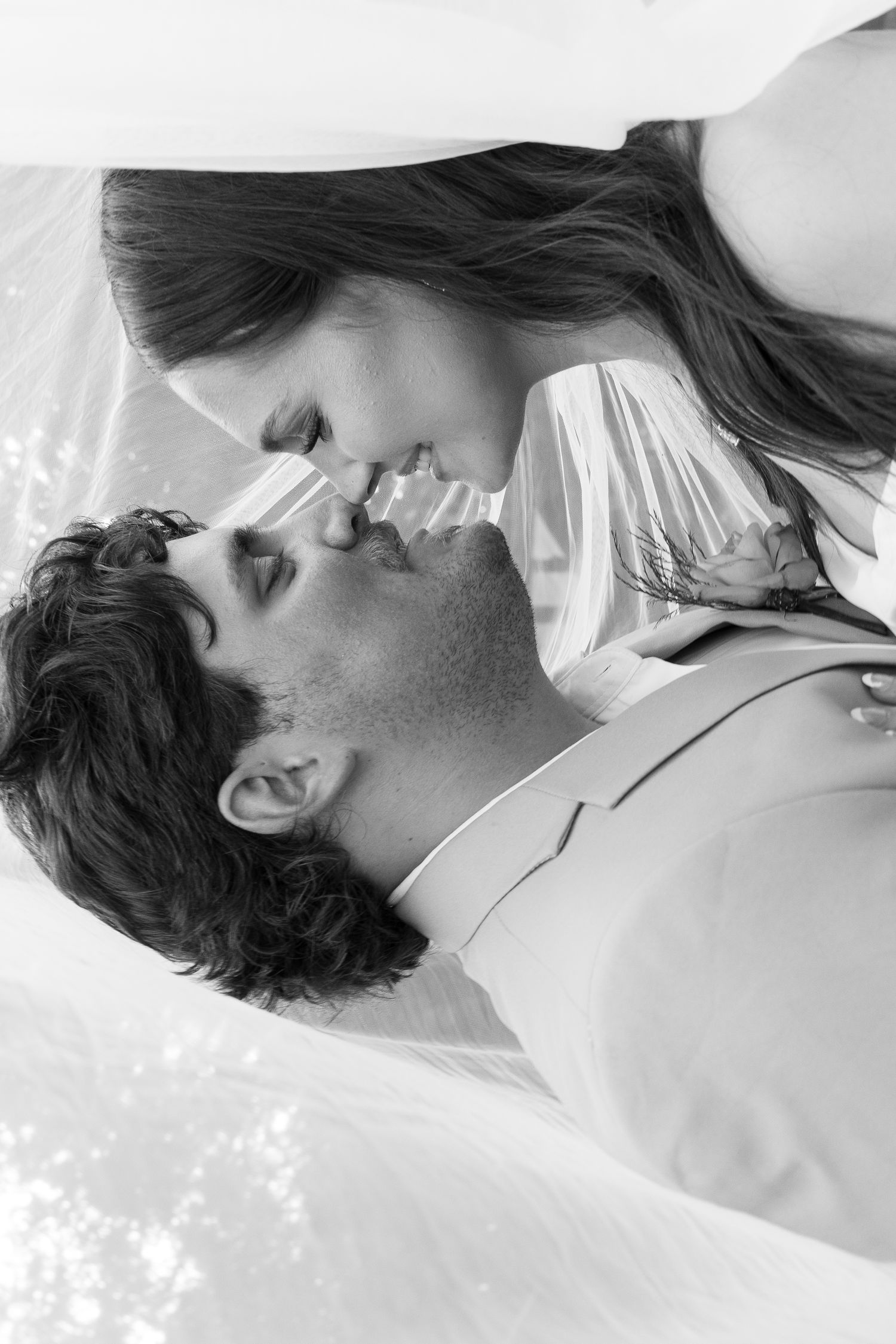 Romantic black and white close-up of bride and groom leaning in for a kiss beneath a flowing white veil outdoors.