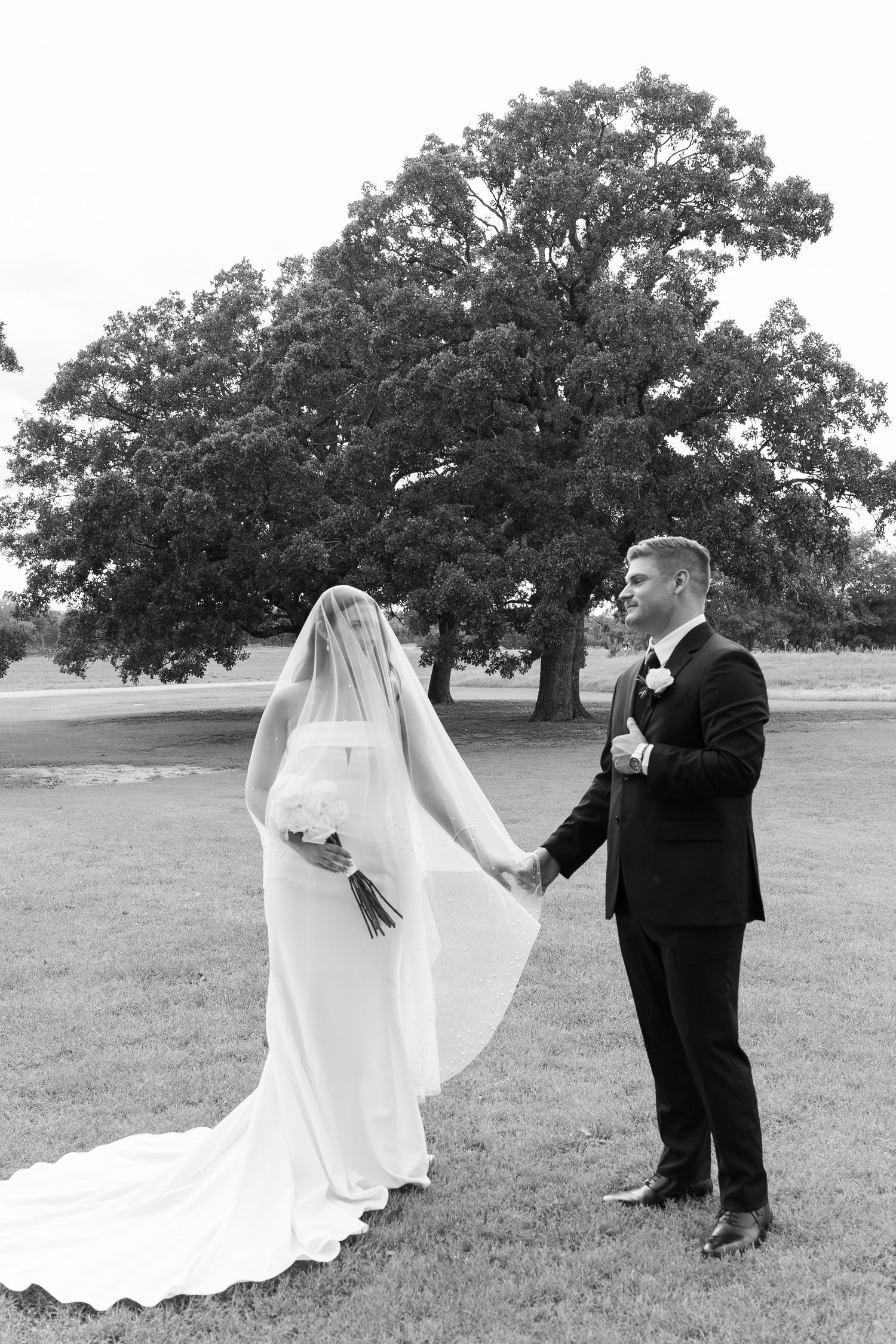 Black and white first look photo of groom holding brides hand as veil covers her face outdoors.