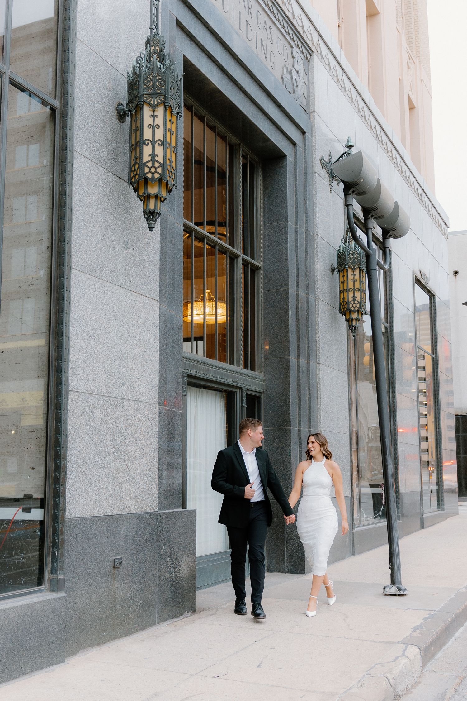 Engaged couple holding hands walking past ornate building exterior during urban engagement session.