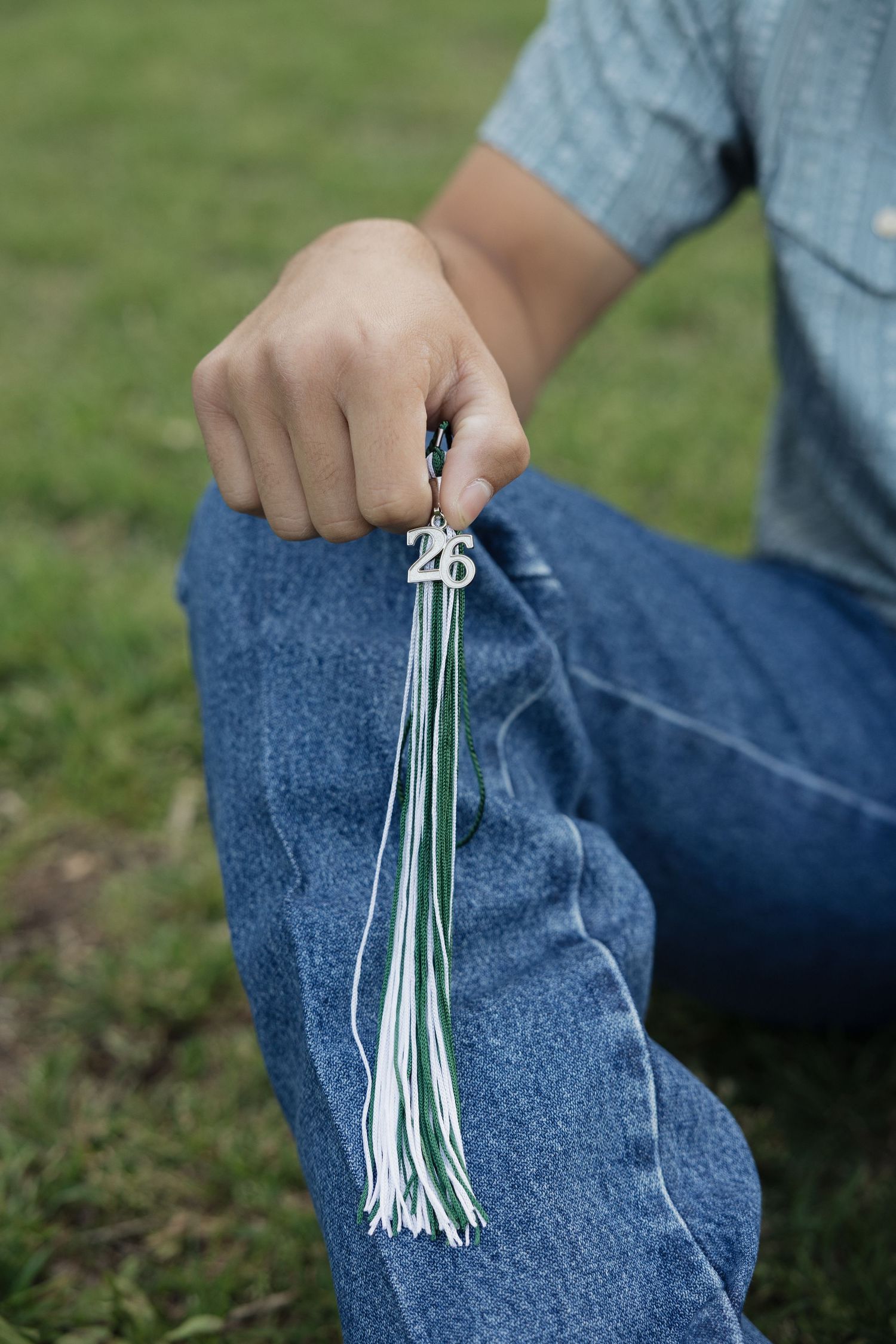 A person in jeans holds a green and white graduation tassel with a 26 charm while sitting on grass.
