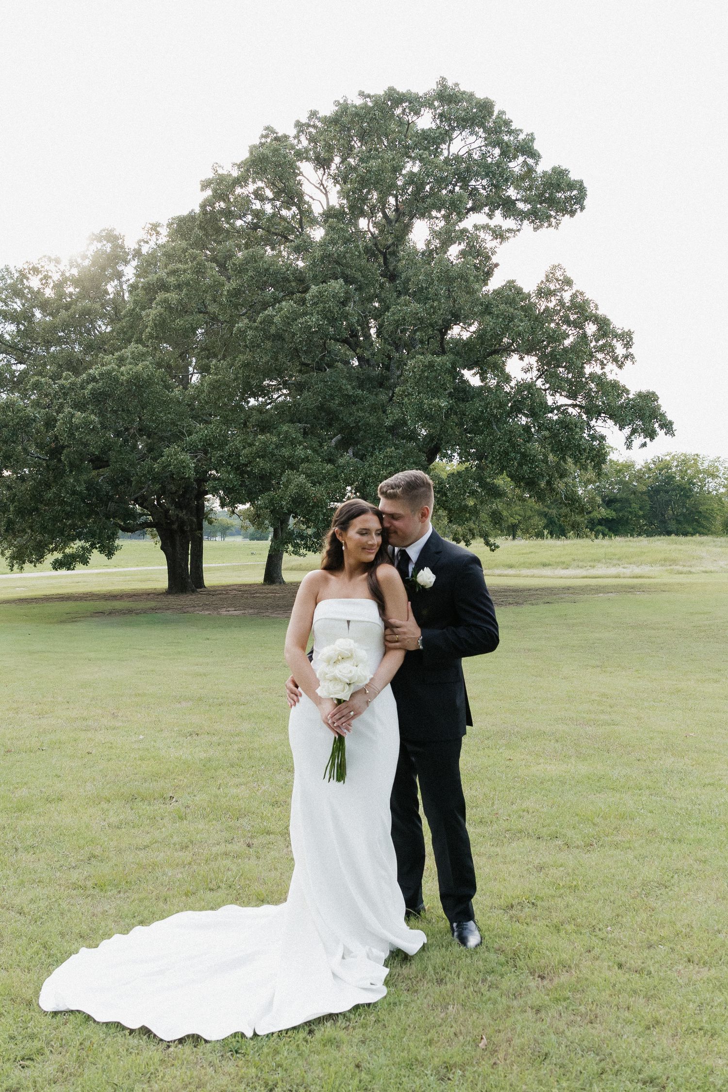 Bride and groom pose together on green lawn with large oak tree backdrop after wedding ceremony.