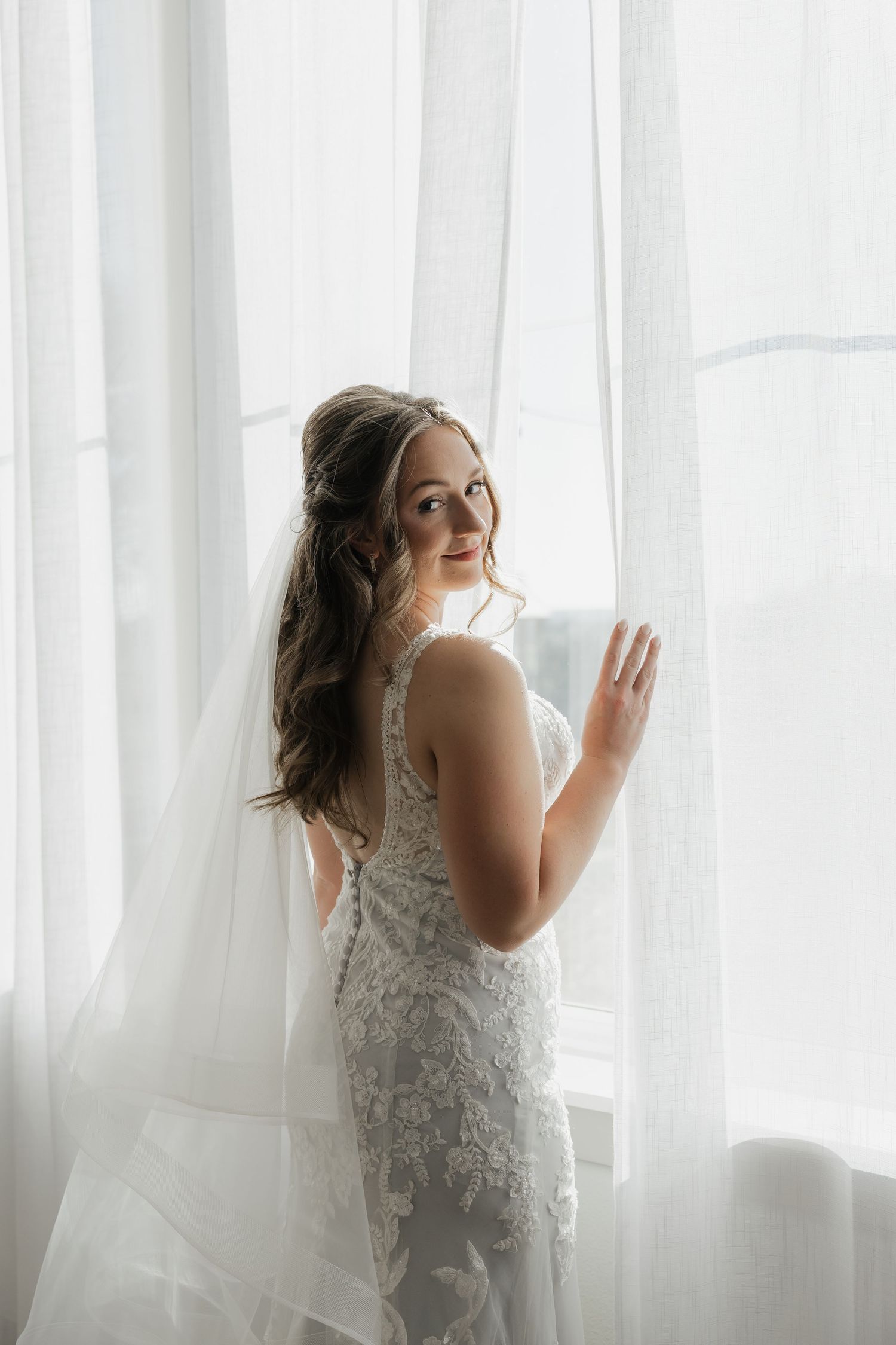 Bride in lace gown and veil smiling while touching sheer curtain by window.