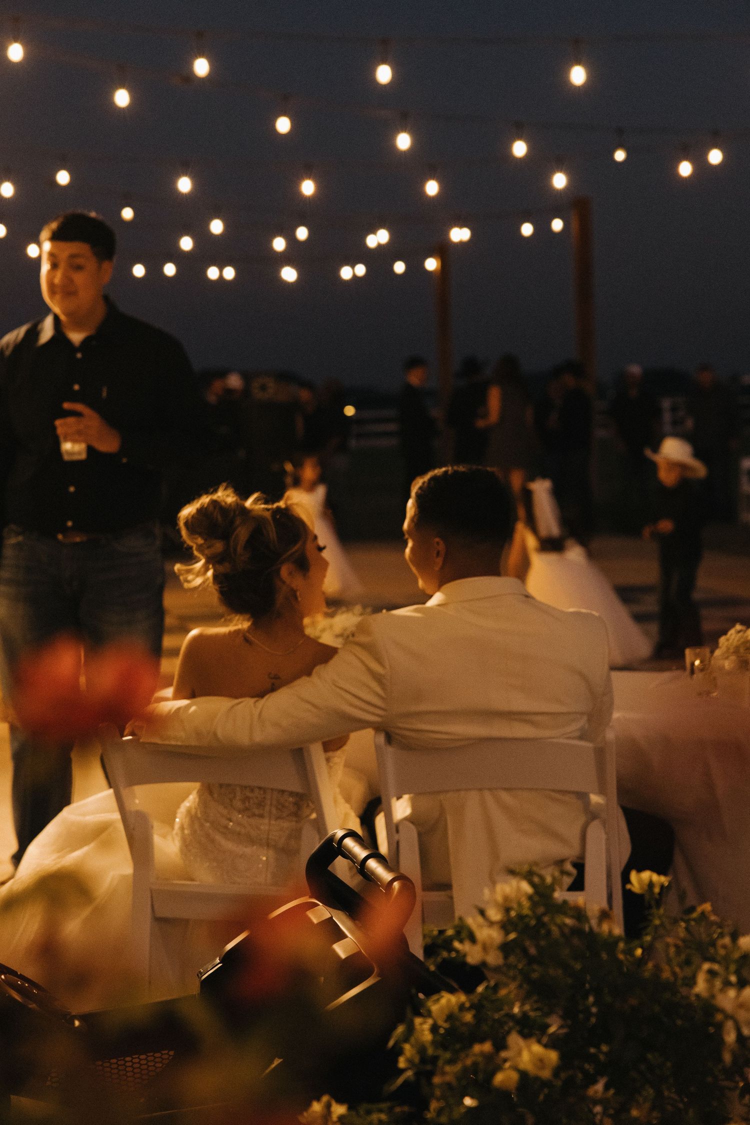 Bride and groom seated together at outdoor evening wedding reception under warm string lights.