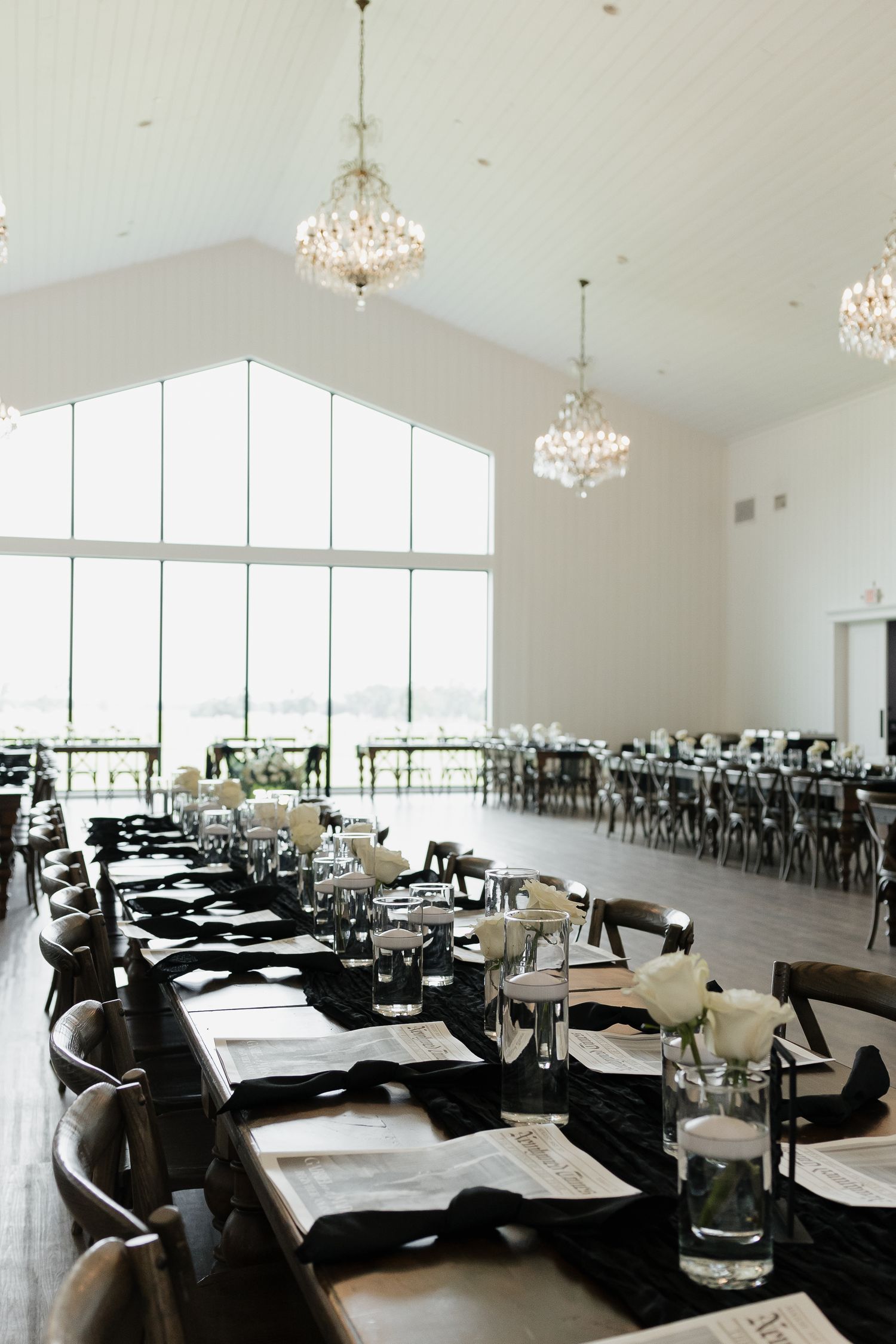 Elegant wedding reception hall with crystal chandeliers, long wooden tables, black runners, and white floral centerpieces.