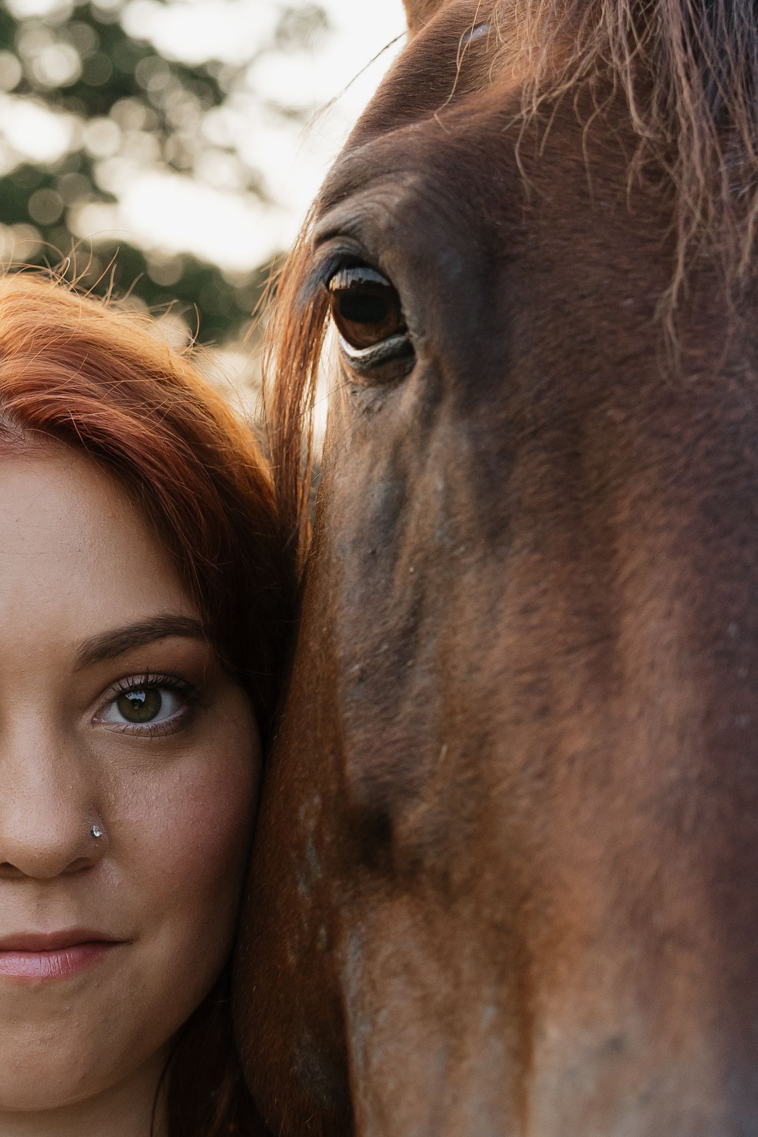 Close-up portrait of a red-haired woman with a nose ring posing cheek-to-cheek with a brown horse.