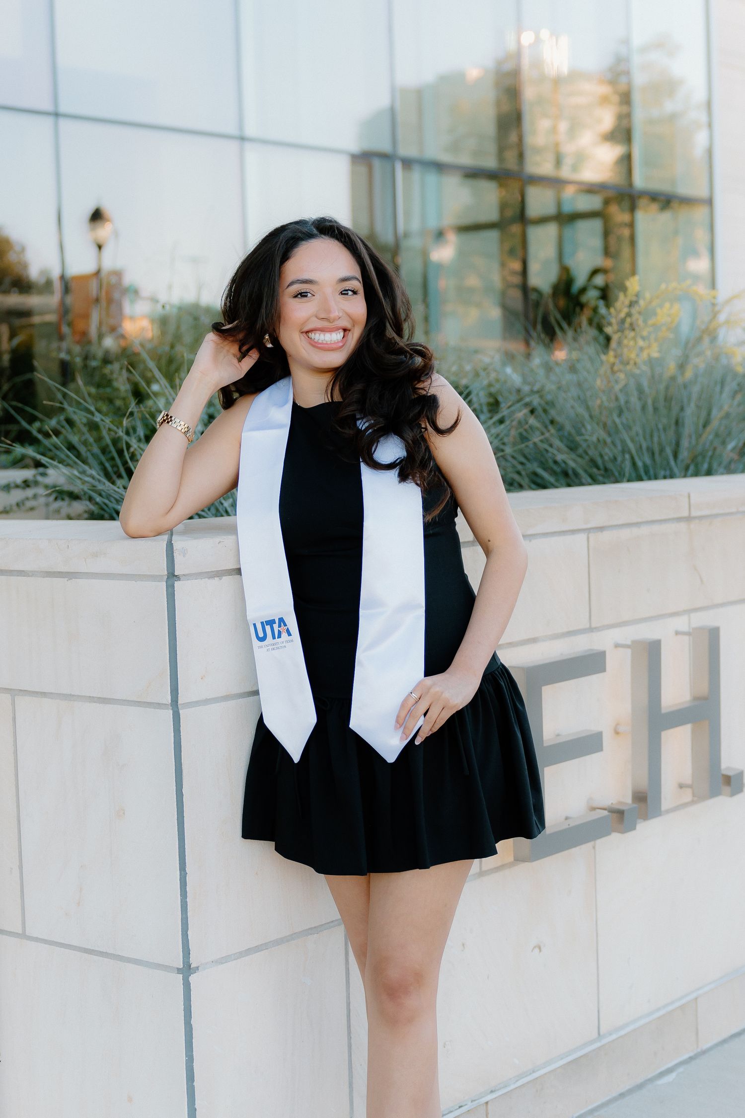 Smiling young woman in black dress with UTA graduation stole posing outdoors.