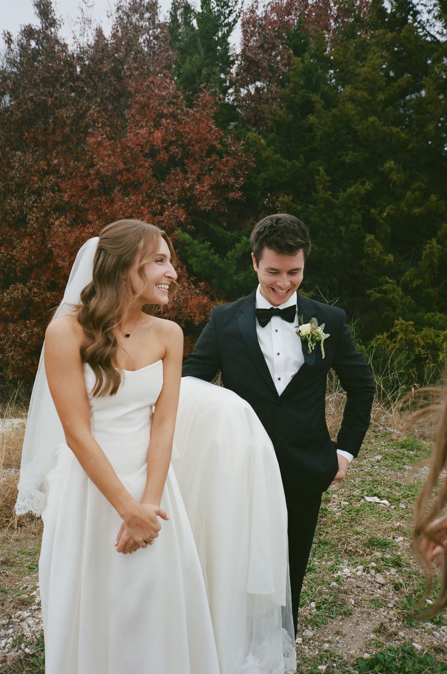 Laughing bride in white gown and groom in black tuxedo among autumn trees.