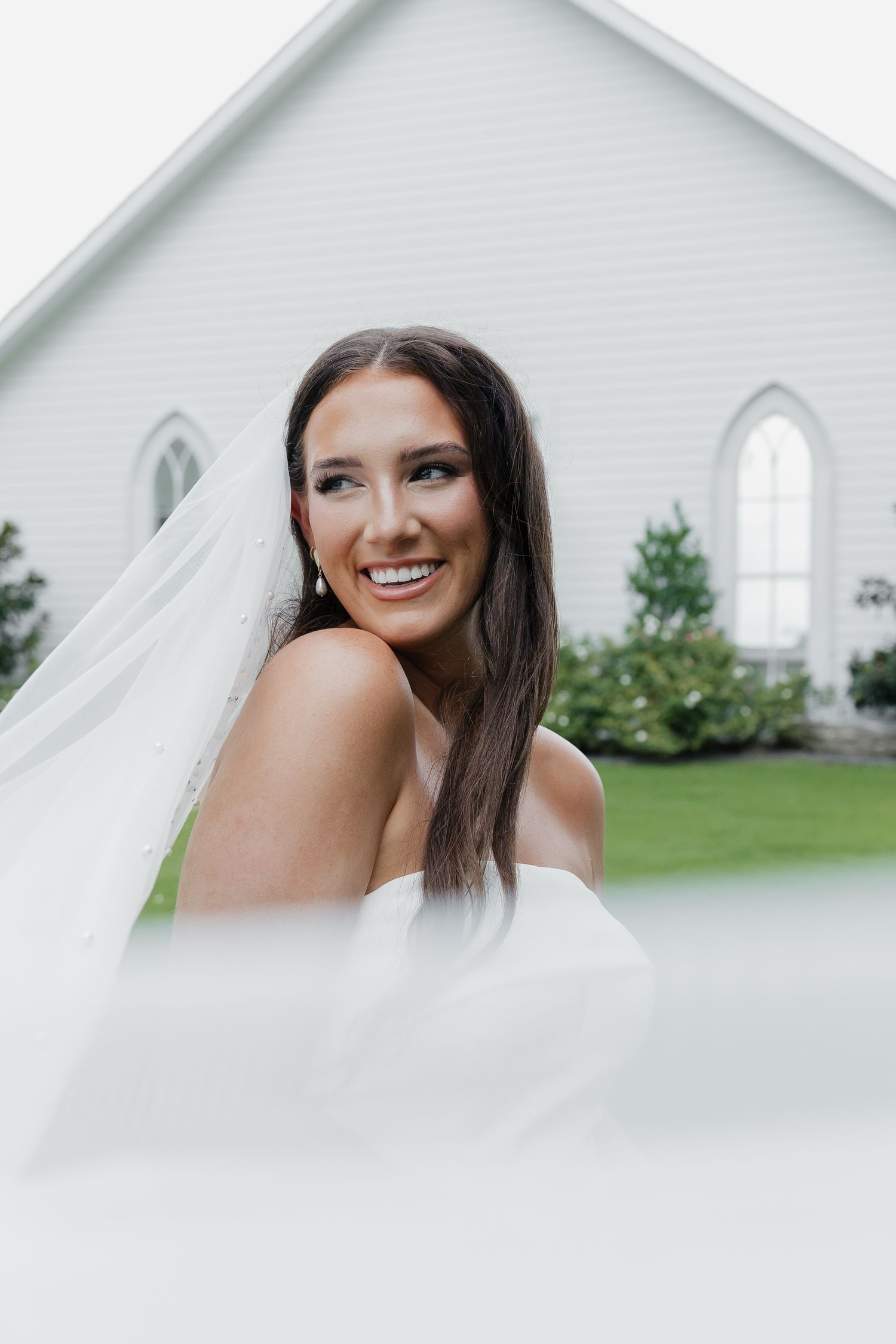 Smiling bride in strapless white gown with flowing veil poses in front of a white chapel.