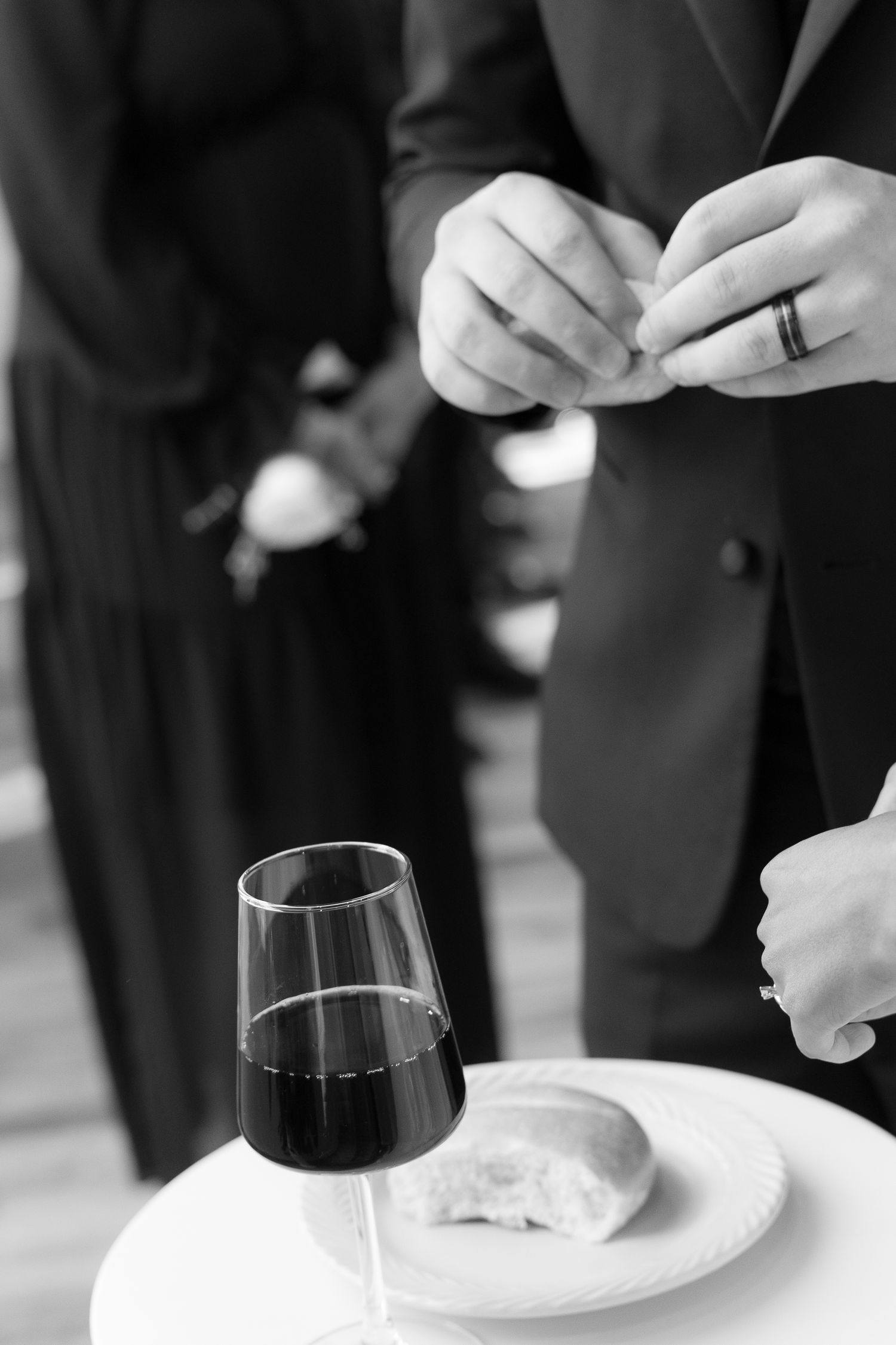 Black and white photo of groom adjusting cufflinks beside wine glass and bread roll on white plate.