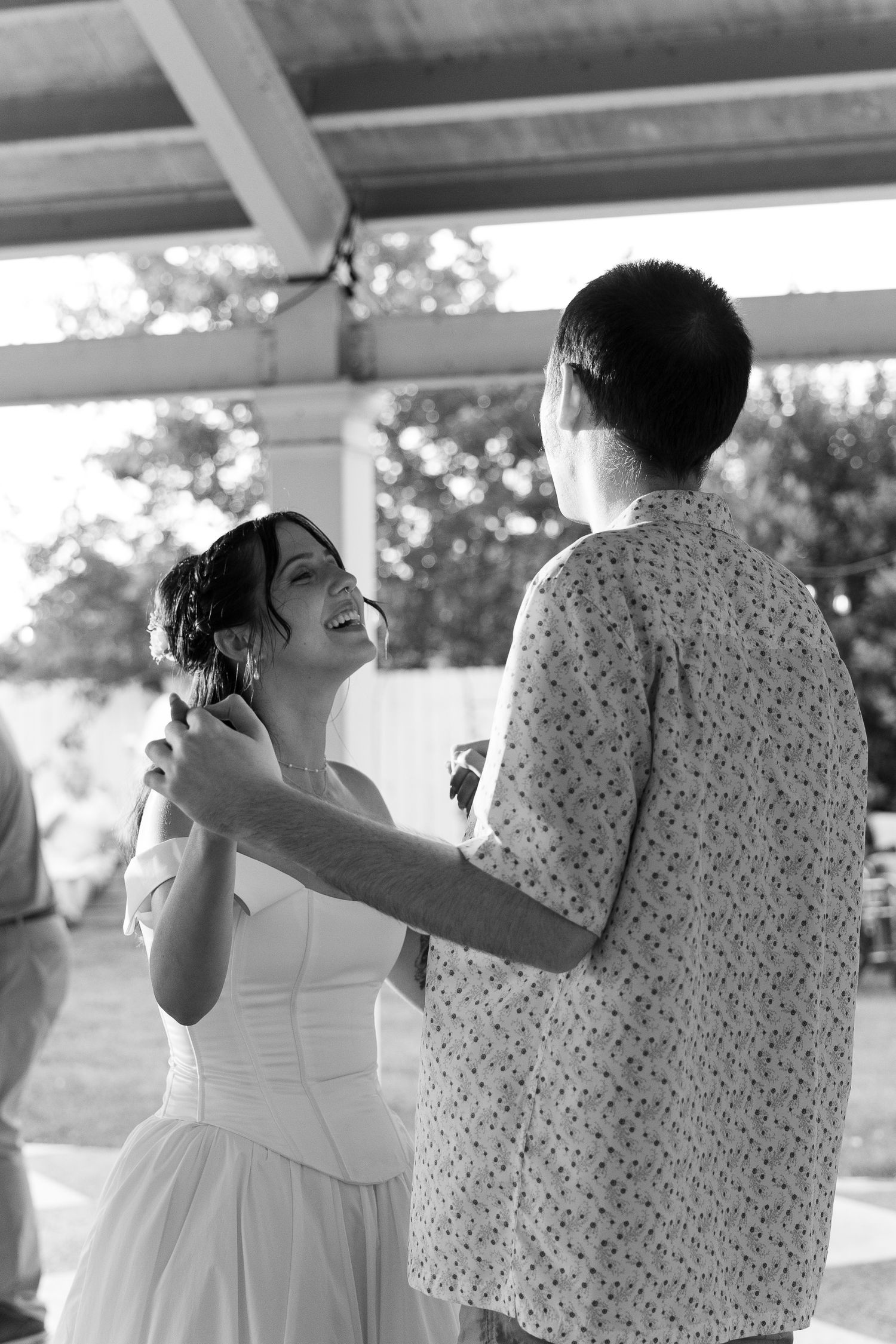 Black and white photo of smiling bride in white dress dancing with a guest under an outdoor covered patio.