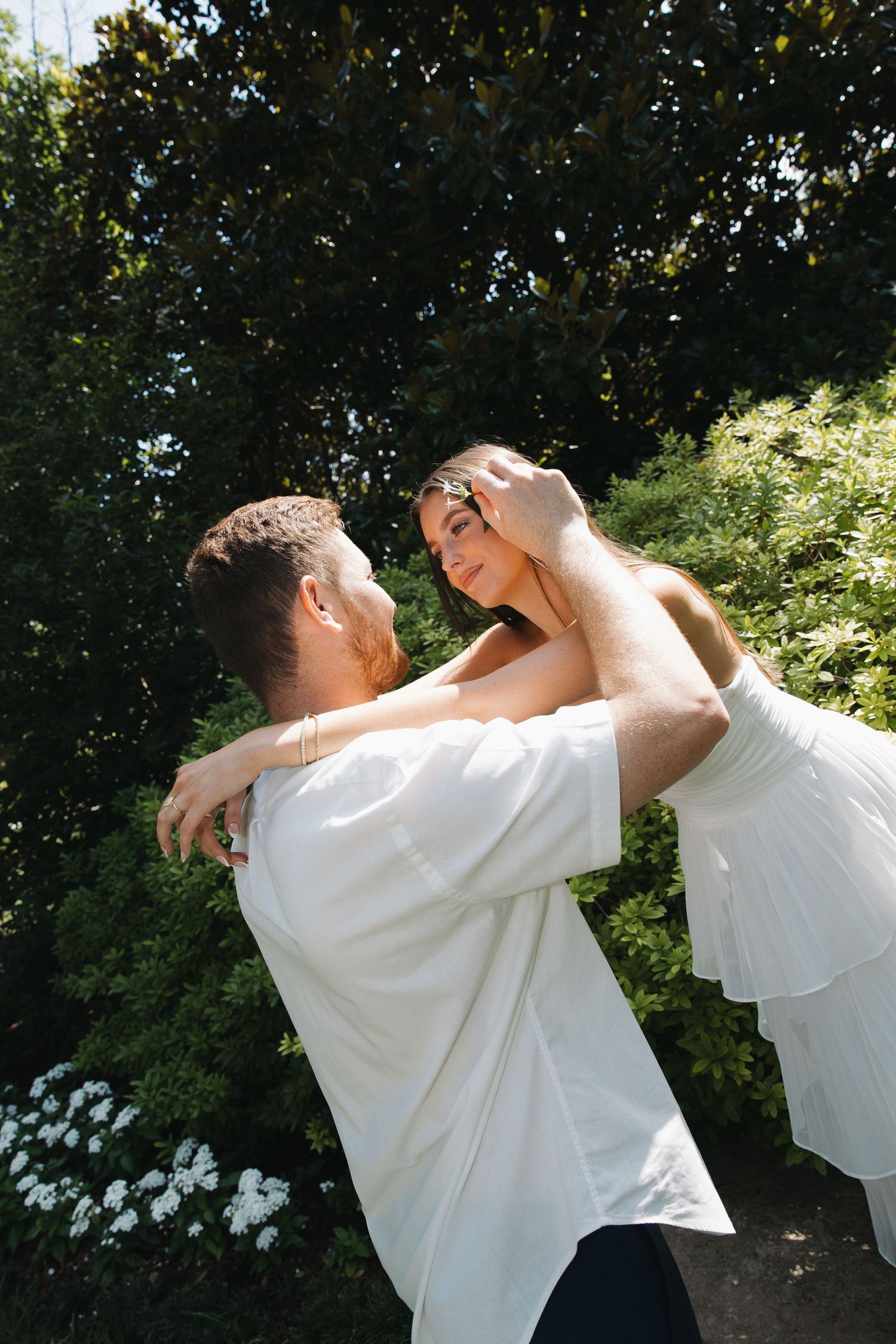 Couple in white outfits dancing together outdoors, surrounded by lush green trees and white flowers.