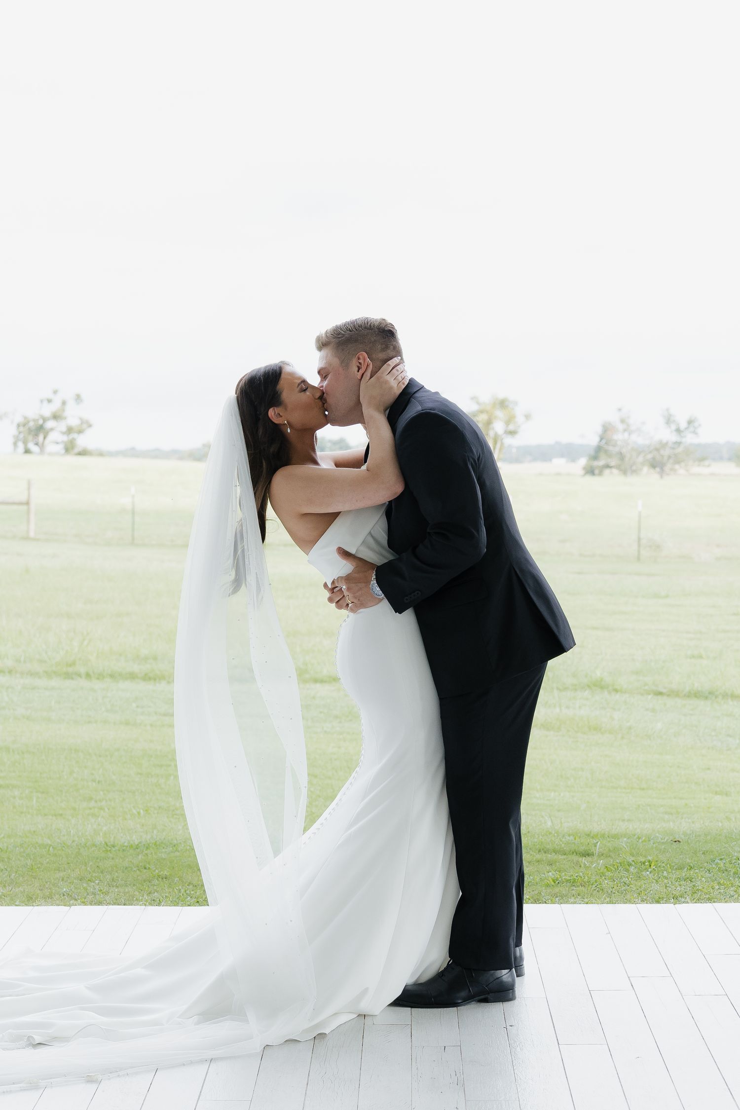 Bride and groom share a romantic kiss on white porch with sweeping green countryside backdrop.