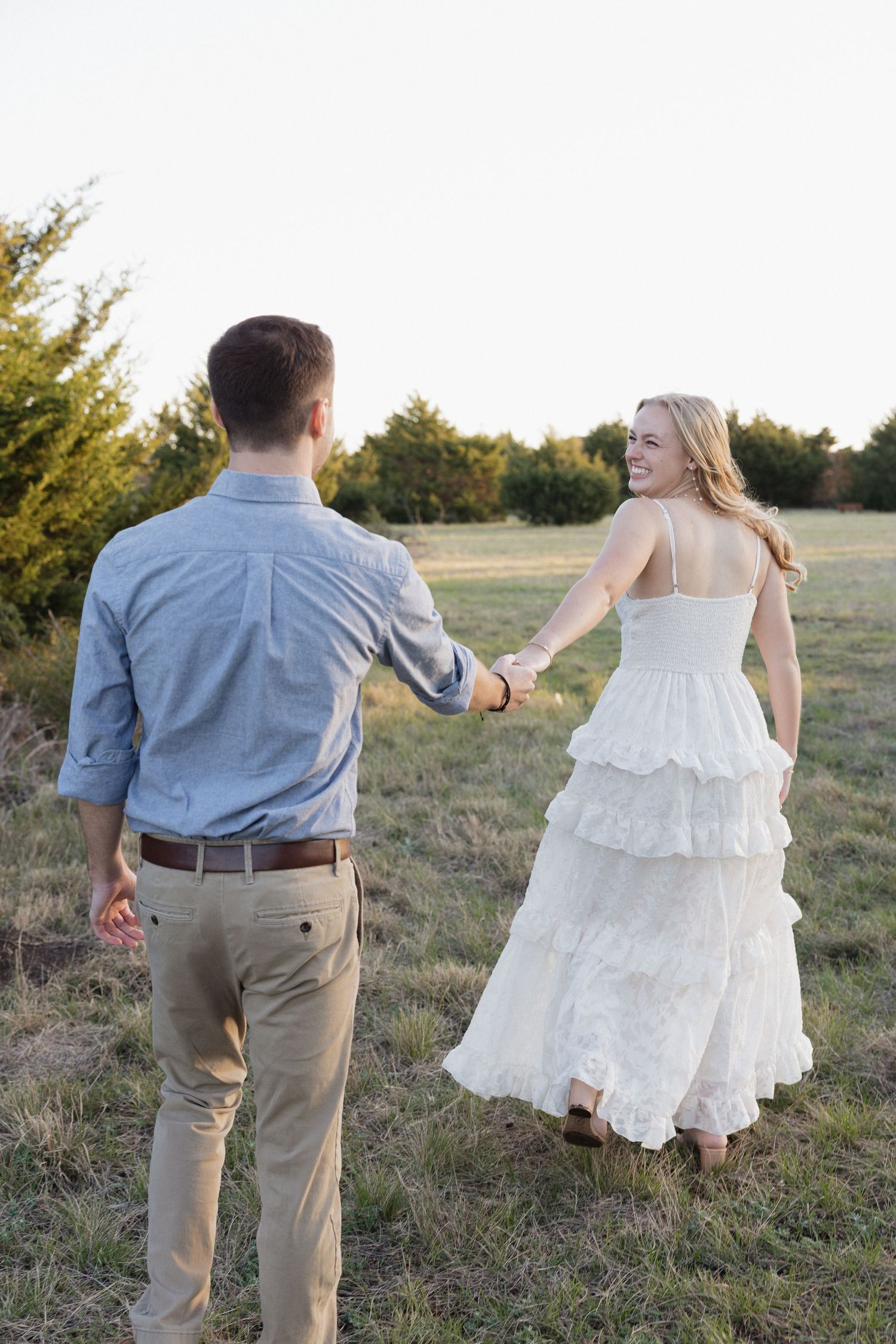A smiling bride in a white ruffled dress holds hands with a man in a blue shirt as they walk through a sunny field.