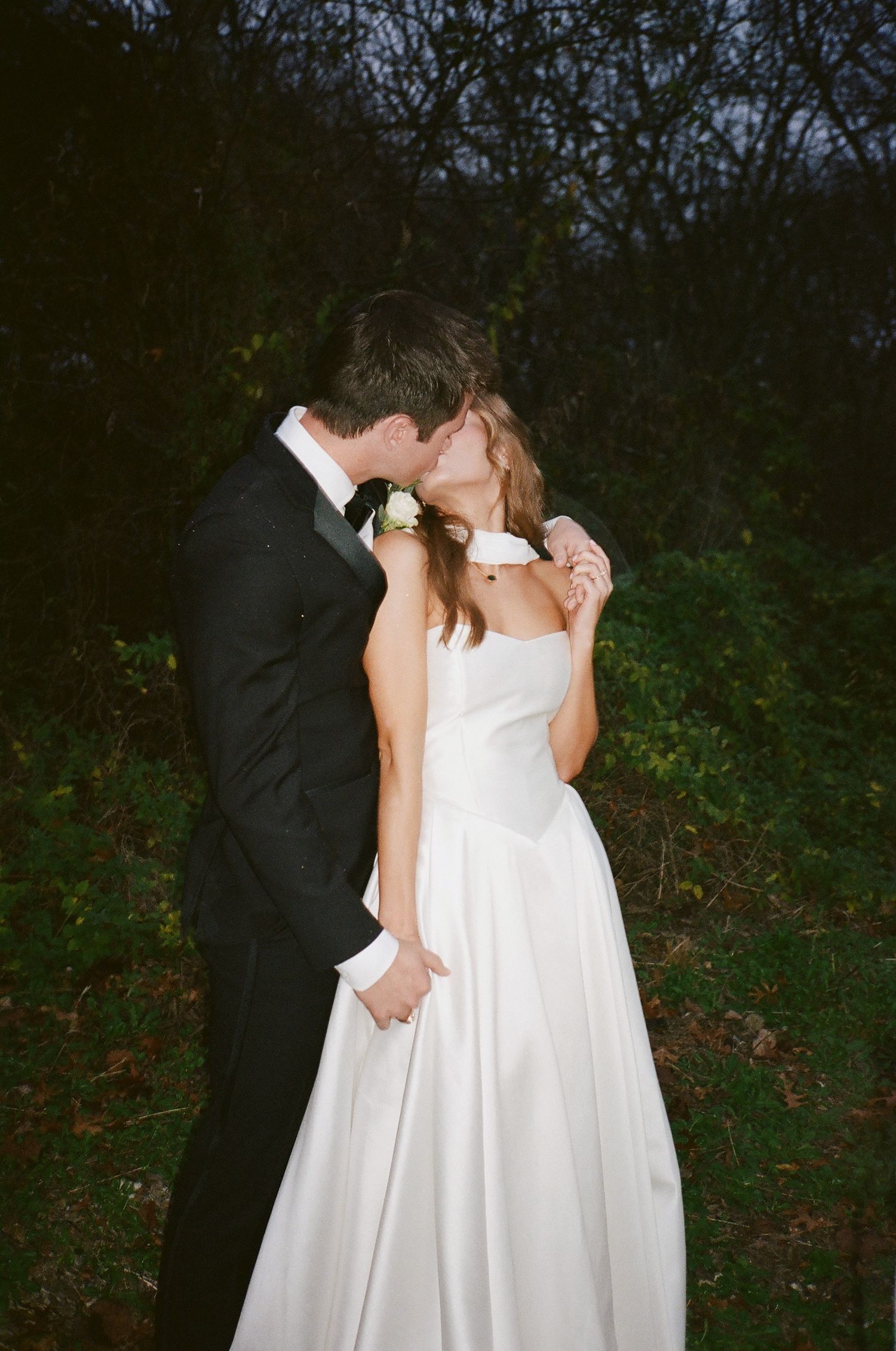 Bride and groom sharing a romantic kiss outdoors at dusk surrounded by dark foliage.
