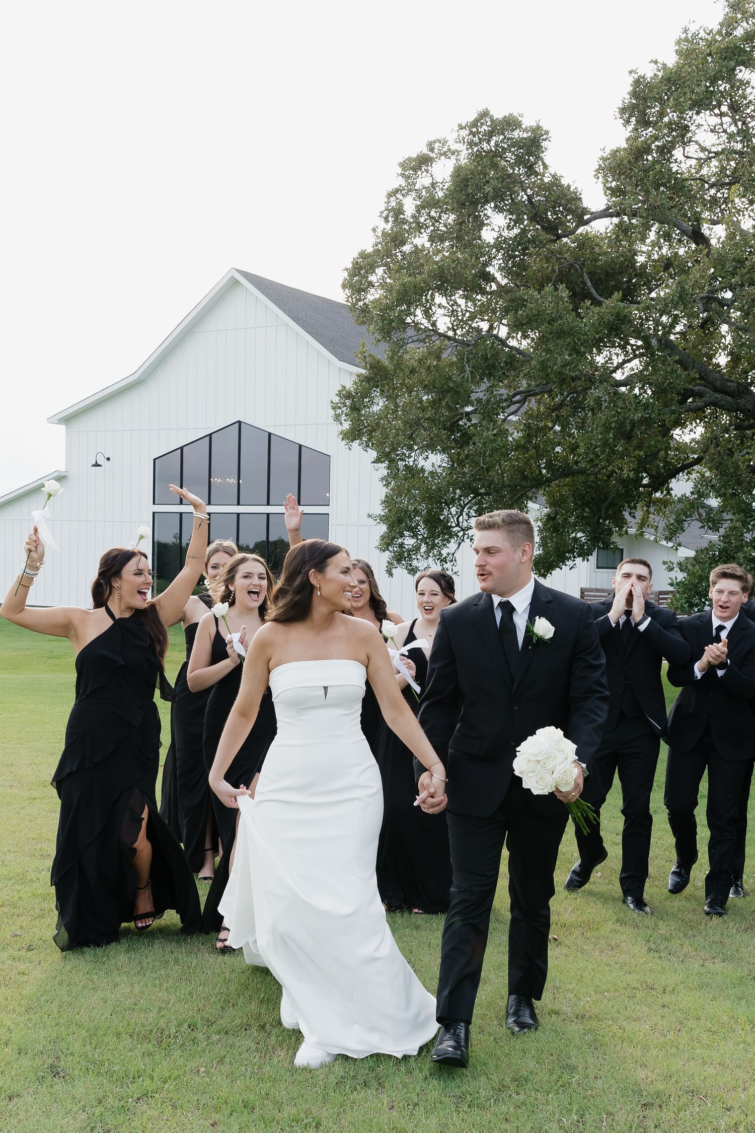 Bride and groom walk hand in hand as wedding party celebrates behind them near white barn venue.