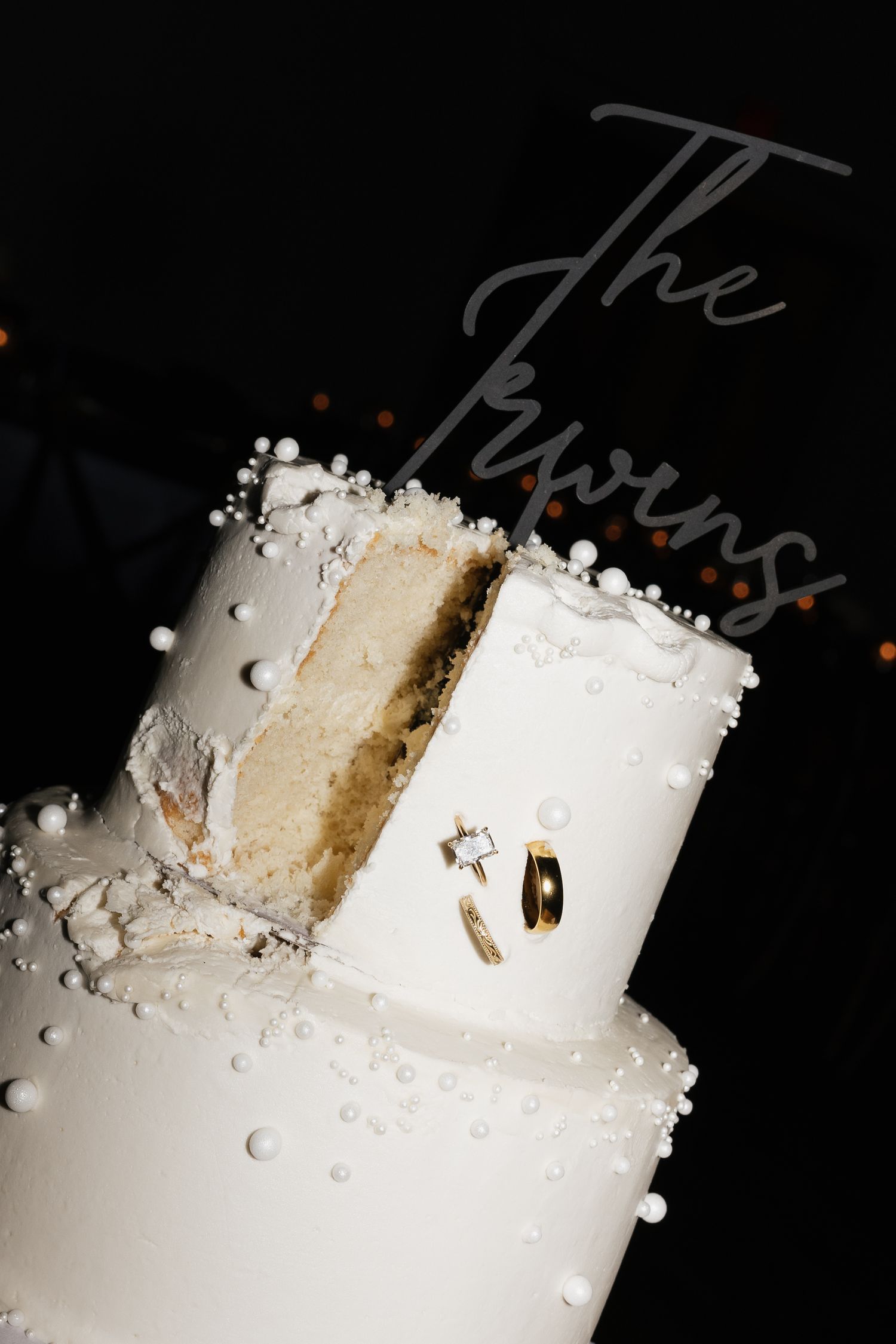White tiered wedding cake with pearl details, cut slice, and The Twins cake topper with wedding rings.