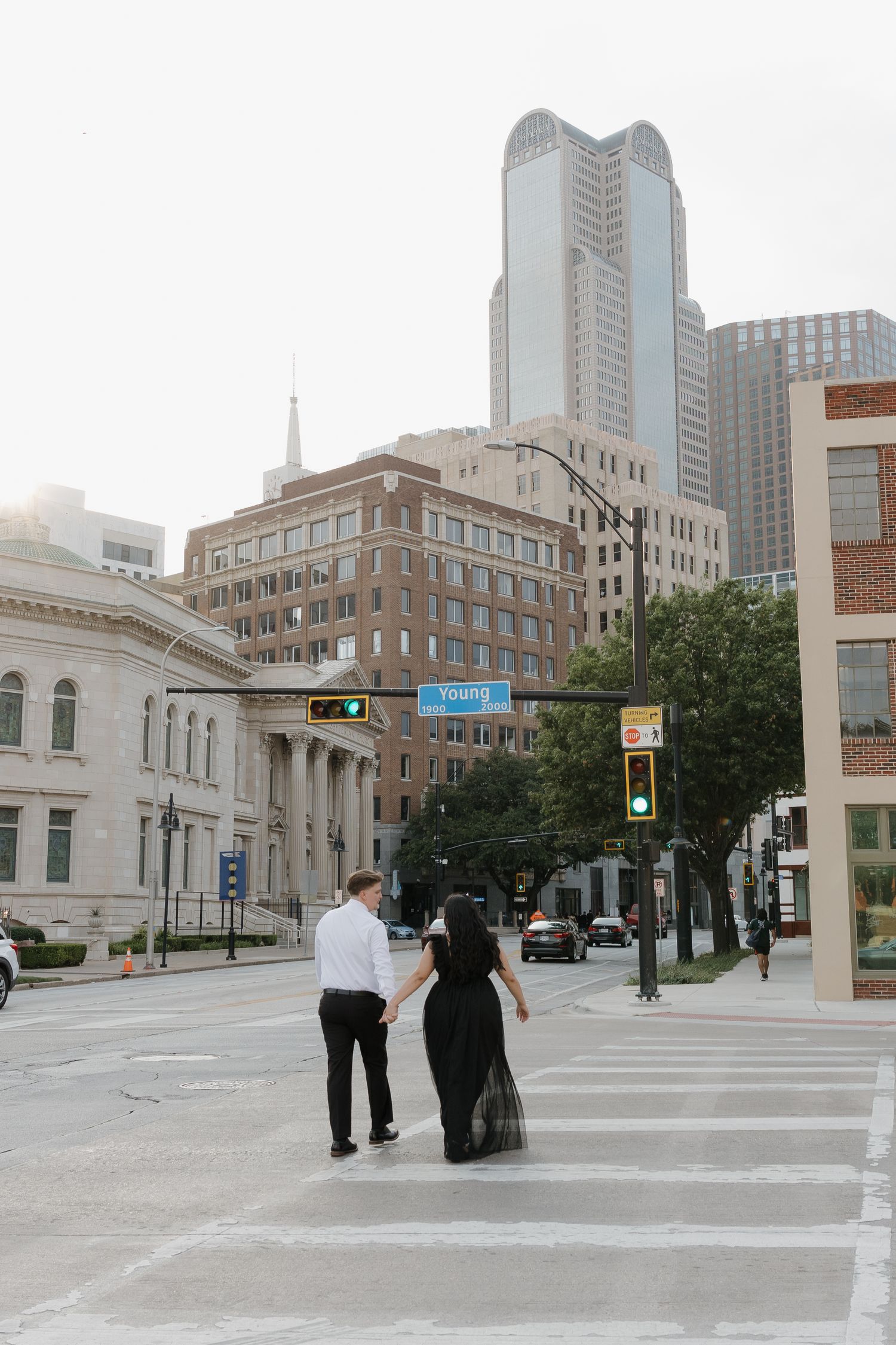 Couple holding hands while crossing a street in downtown Dallas with city skyscrapers in the background.
