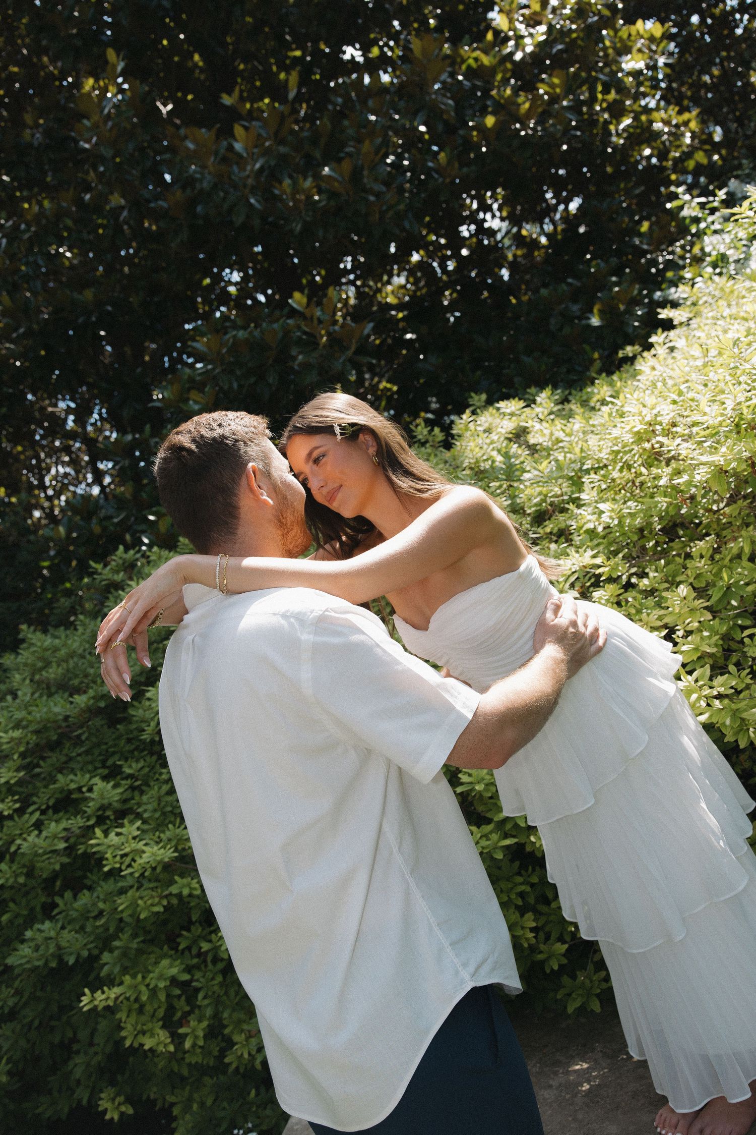 A groom lifts his bride in a white dress outdoors, surrounded by lush green trees on their wedding day.