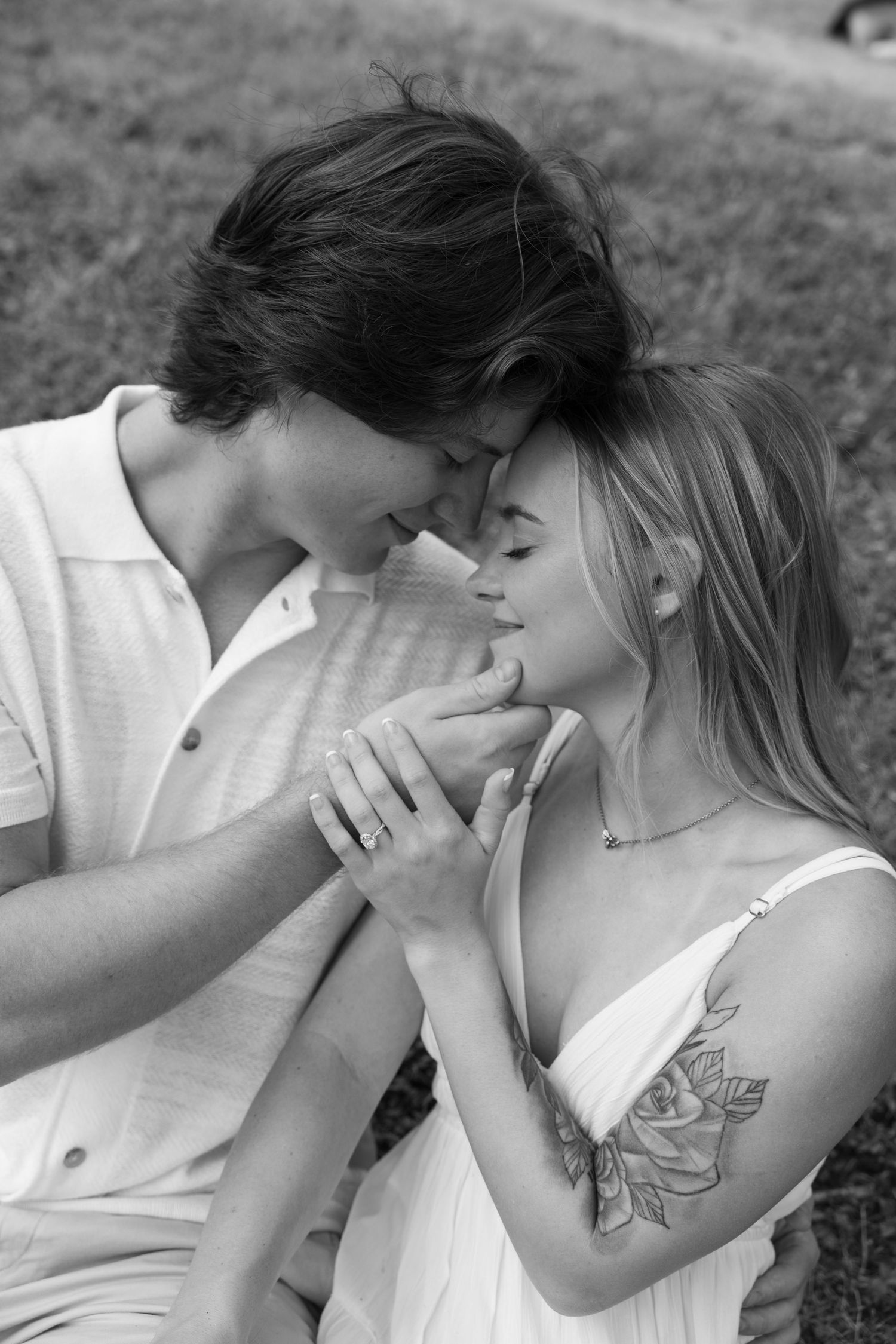Black and white photo of a romantic couple touching foreheads, woman with floral tattoo and engagement ring.
